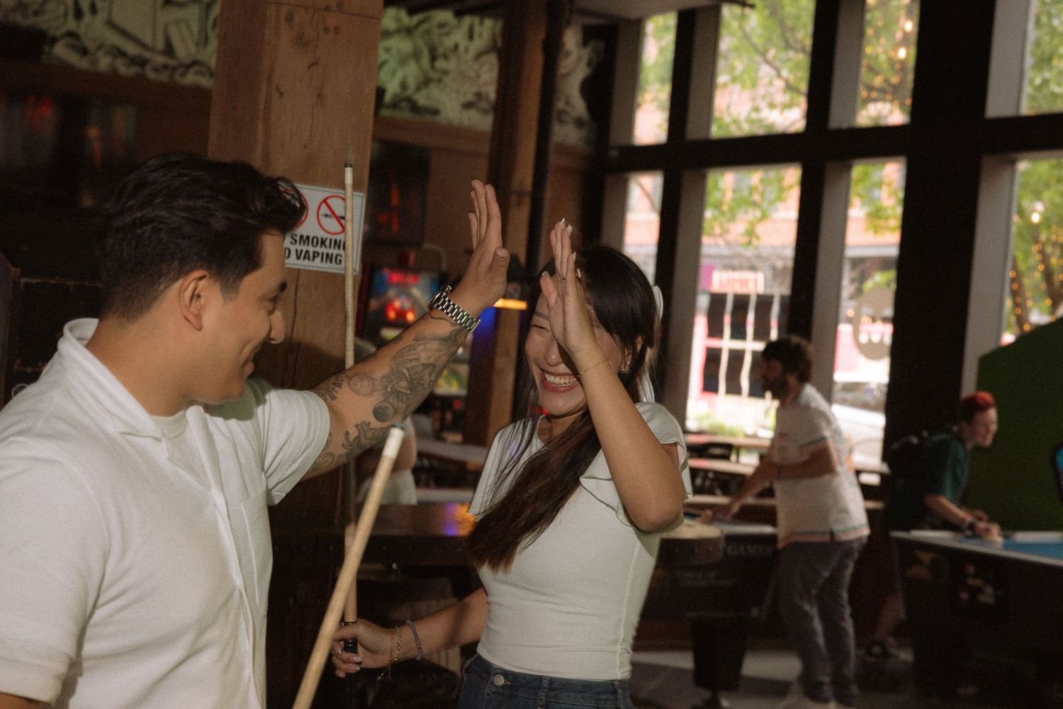 A man and woman high-fiving and laughing near a pool table in a casual bar setting.