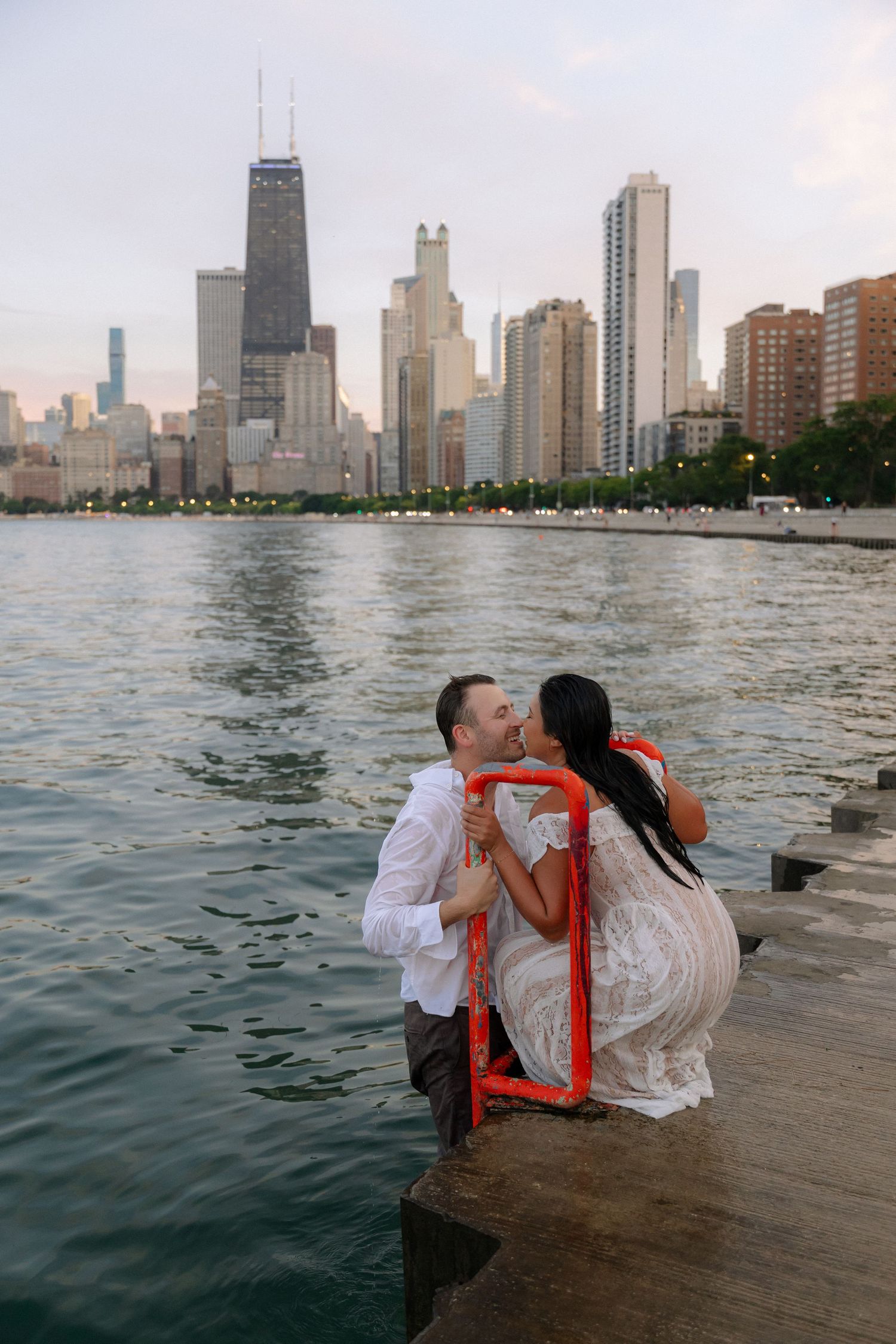 Couple kissing on Chicago lakefront pier with city skyline and John Hancock Center in background at dusk.