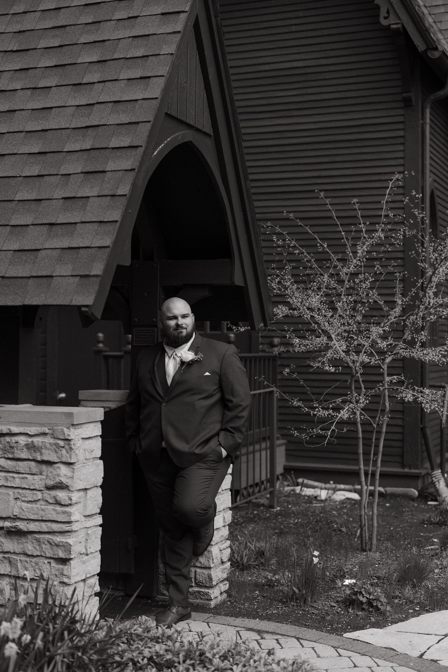 A man in a suit leans against a stone pillar outside a wooden chapel in this black and white portrait photo.