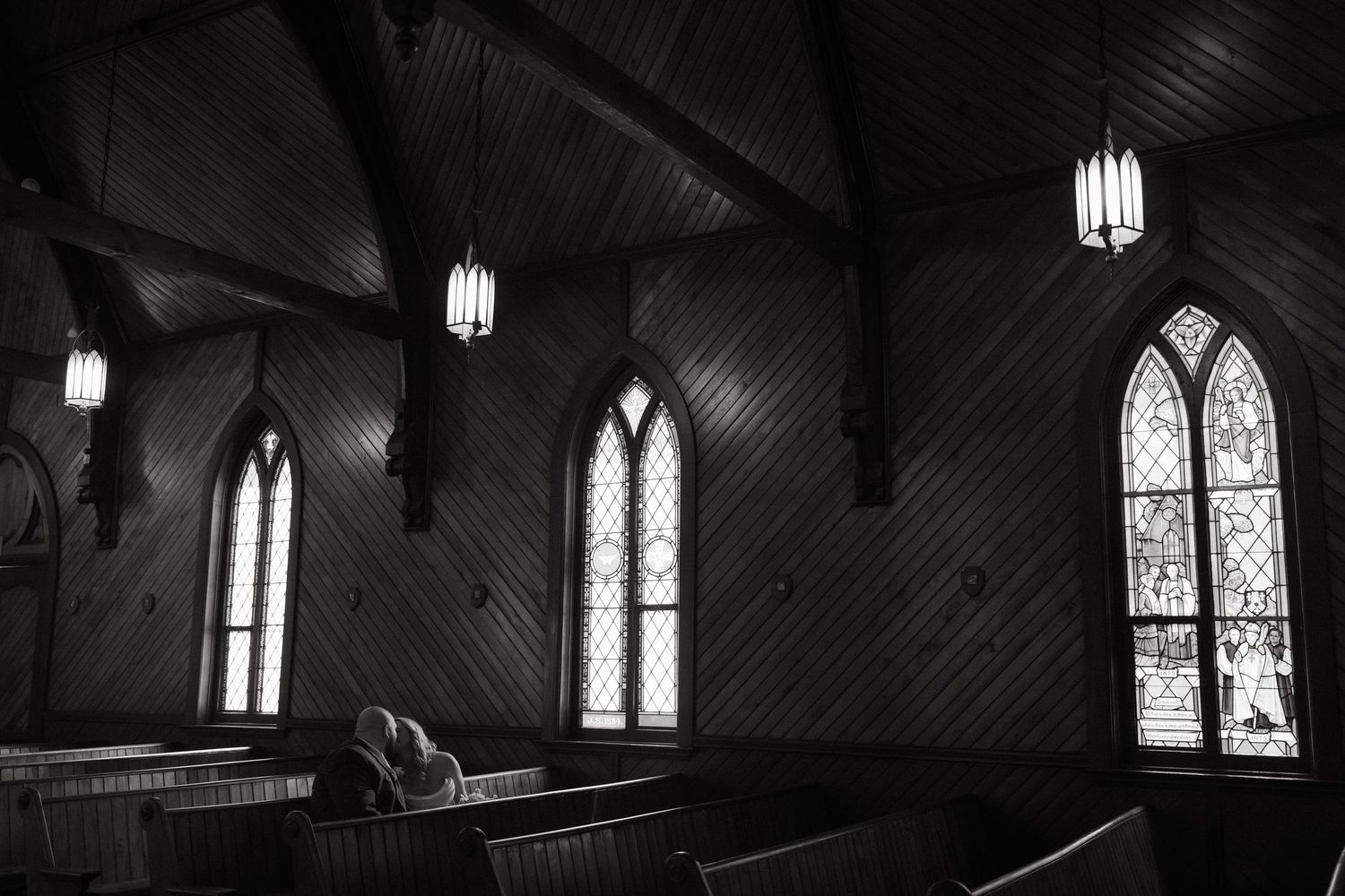 Black and white photo of a person praying alone in a historic wooden church with arched stained glass windows.