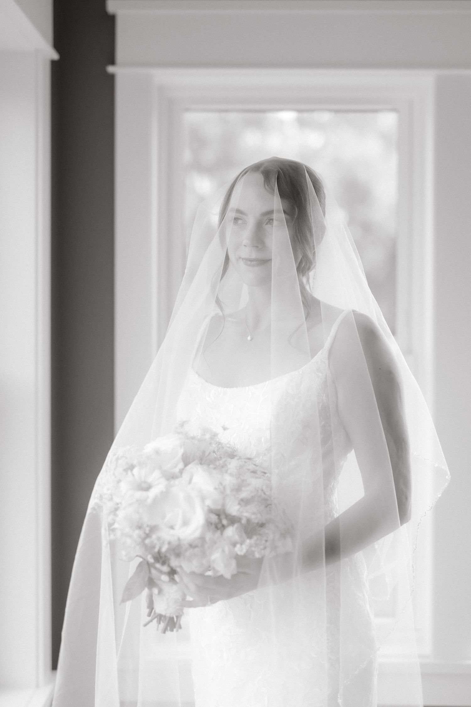 Black and white portrait of serene bride in lace gown and veil holding bouquet, softly lit by window light.