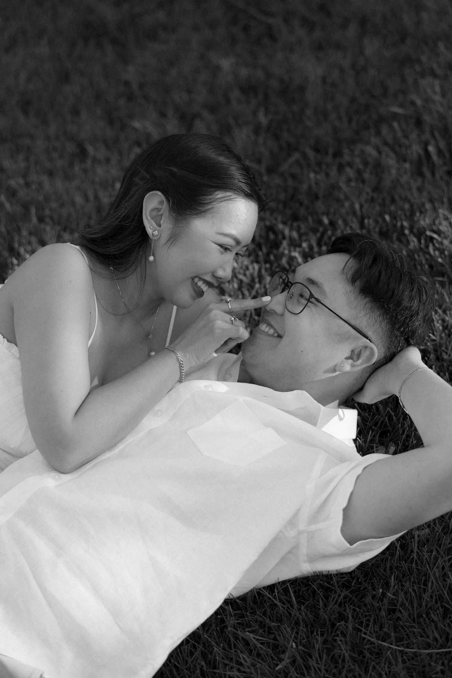 Black and white photo of a joyful couple lying on grass, laughing together in a romantic outdoor setting.