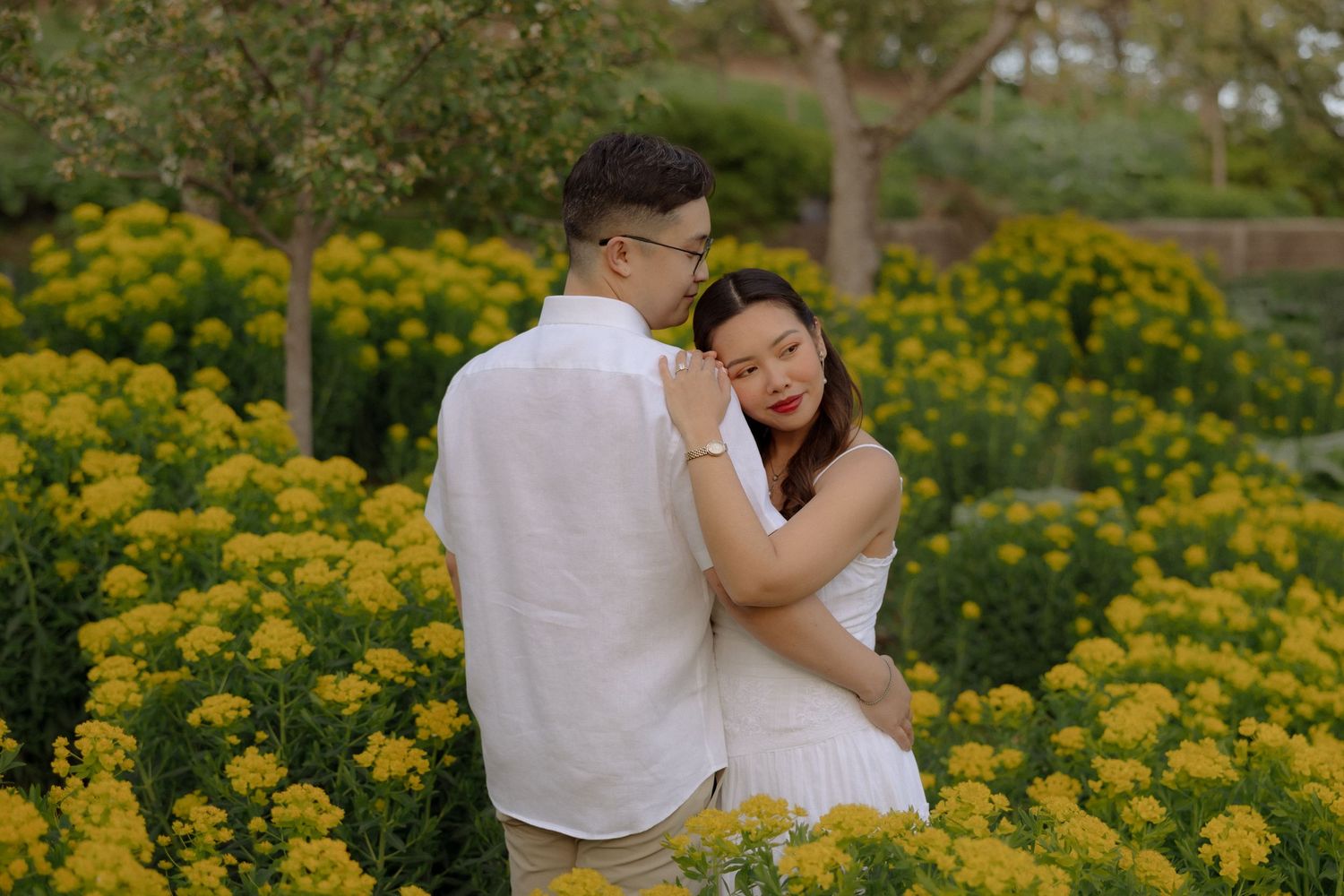 Couple embracing romantically among vibrant yellow flowers in a lush garden, both wearing white outfits.