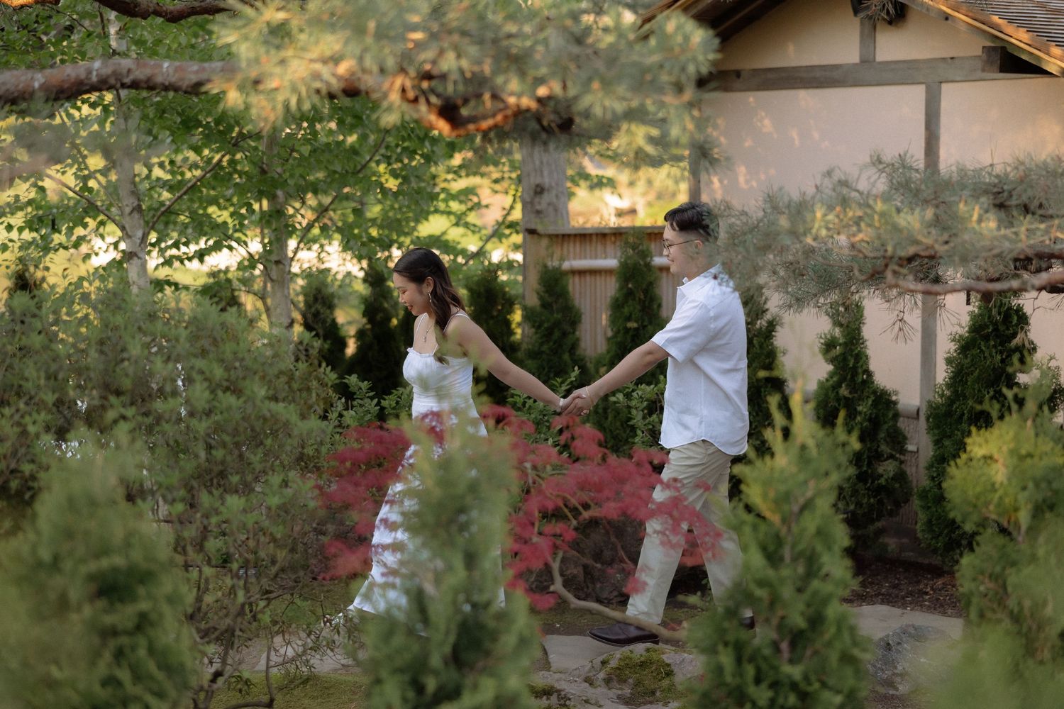 Couple holding hands walking through a lush Japanese garden with evergreen trees and a traditional building.