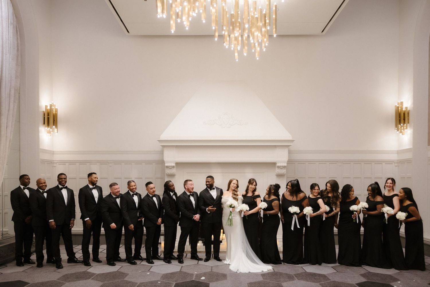 Elegant wedding party in all-black attire poses together in a grand white ballroom with gold chandeliers and sconces.