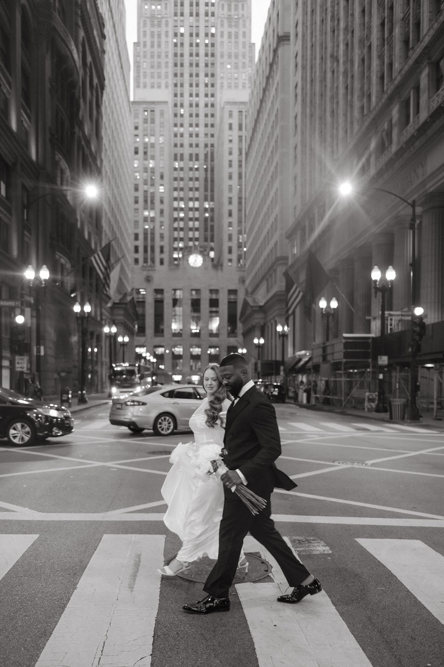 Black and white photo of a bride and groom crossing a city street at night with tall buildings in the background.