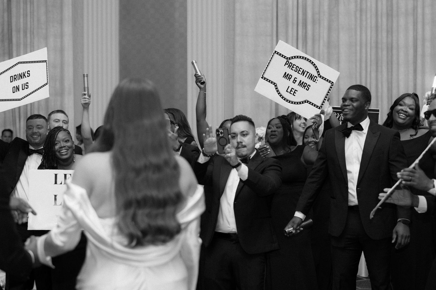 Black and white wedding photo of guests cheering and holding signs at a formal reception celebration.