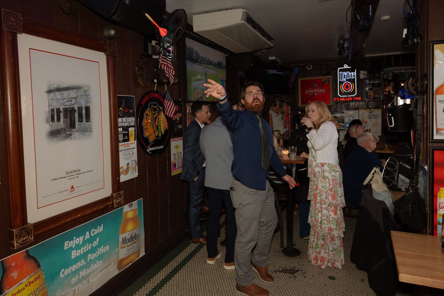 Man taking selfie with woman in a lively bar decorated with sports memorabilia and neon signs.