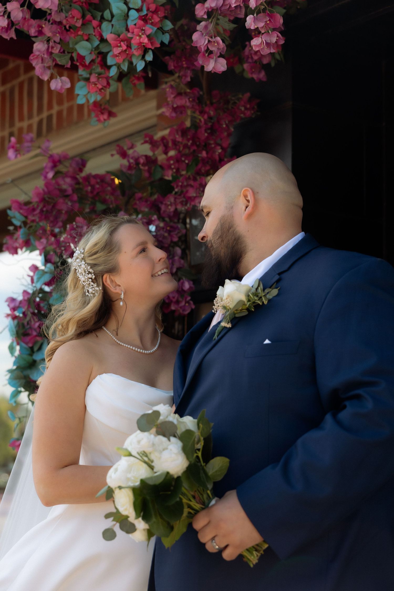 Bride and groom share a loving glance under vibrant pink bougainvillea, bride holding white rose bouquet in strapless gown.