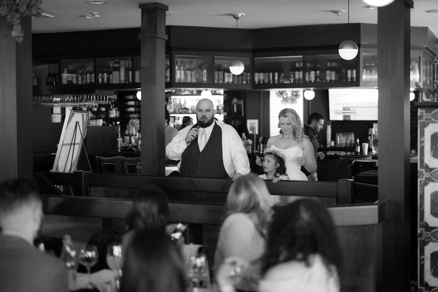 Black and white photo of a groom speaking into a microphone beside his bride at a wedding reception in a restaurant.