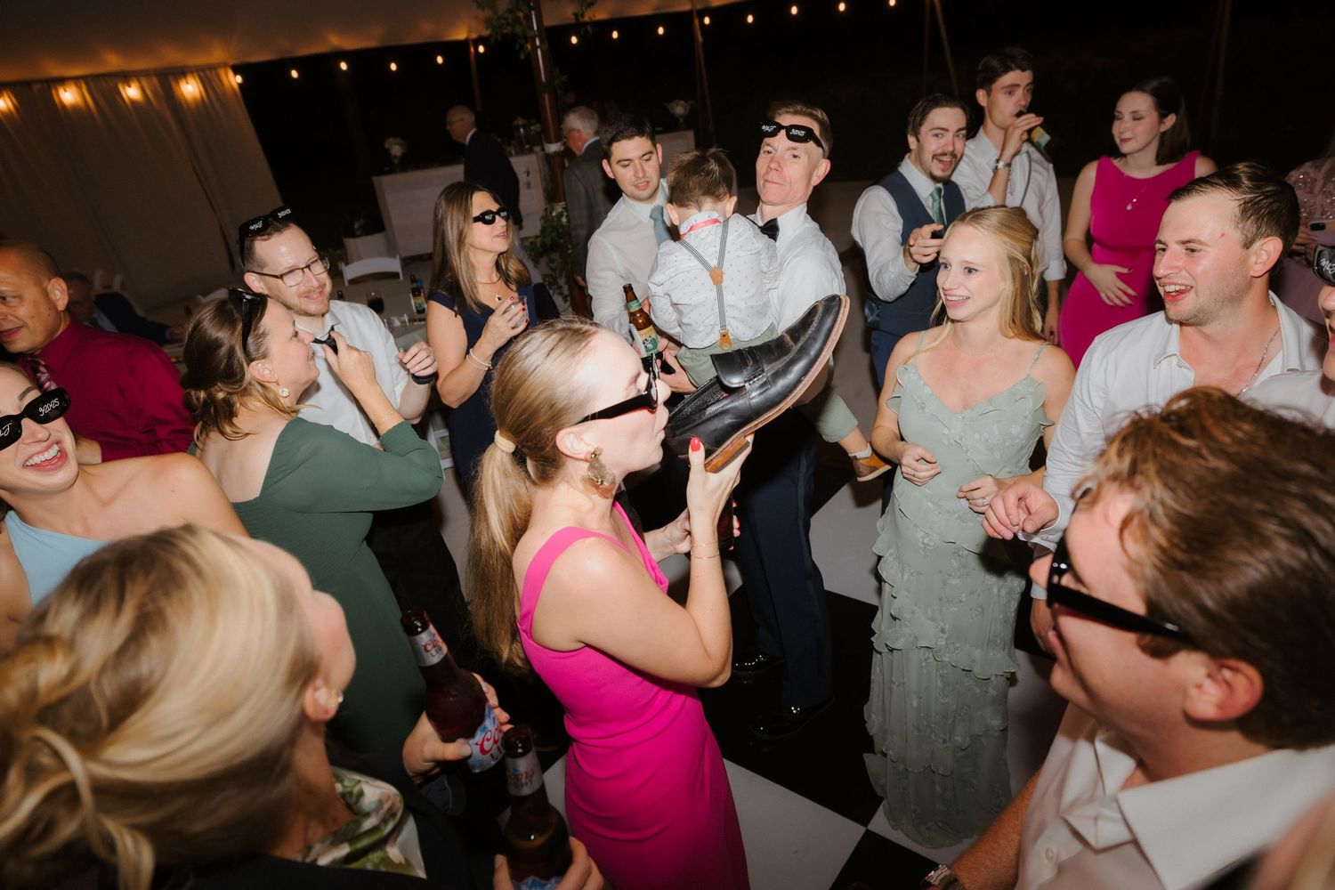 Wedding guest drinks from a shoe while others wear sunglasses and dance at lively wedding reception.