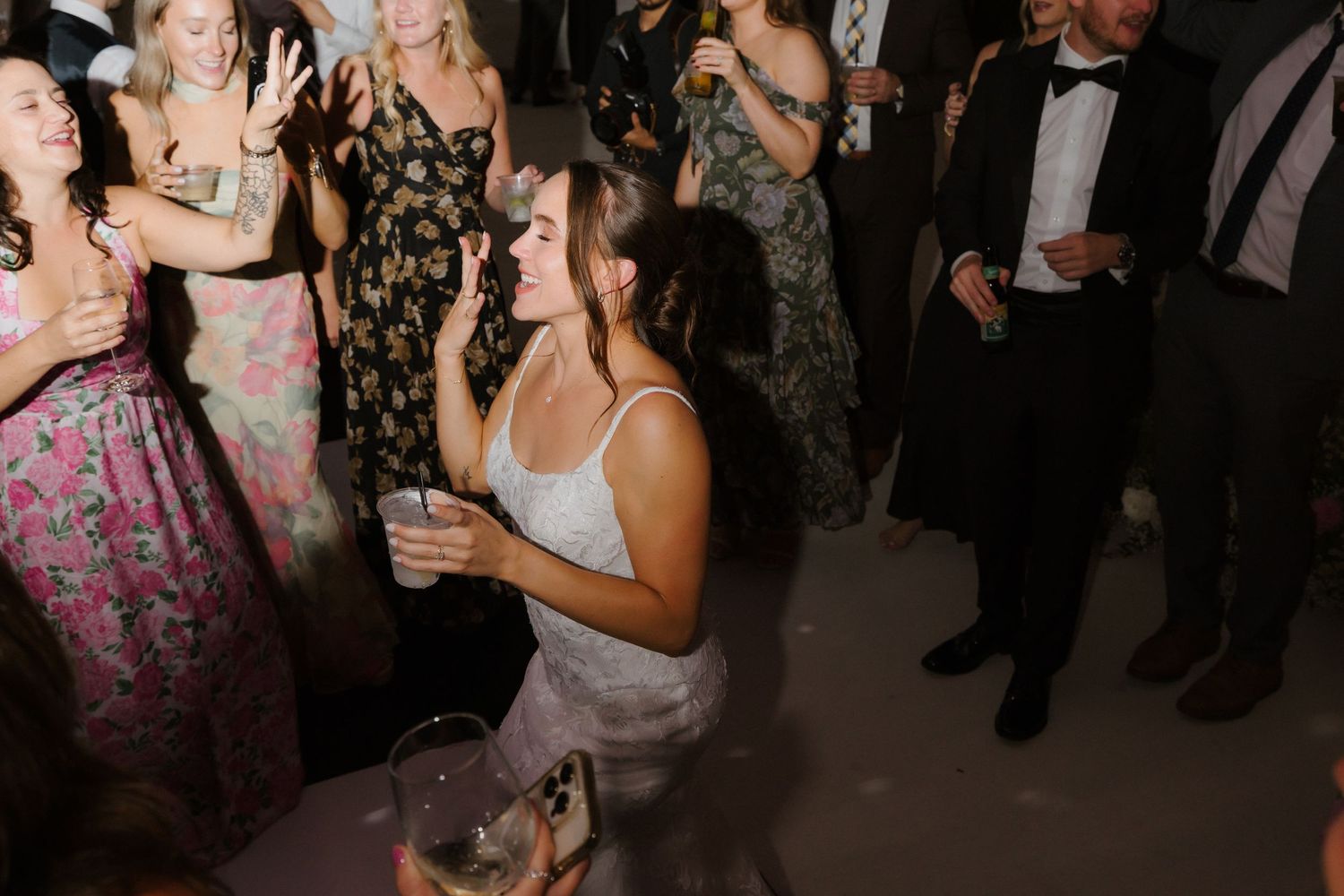 Bride in white gown laughs and dances with wedding guests during a lively reception celebration.