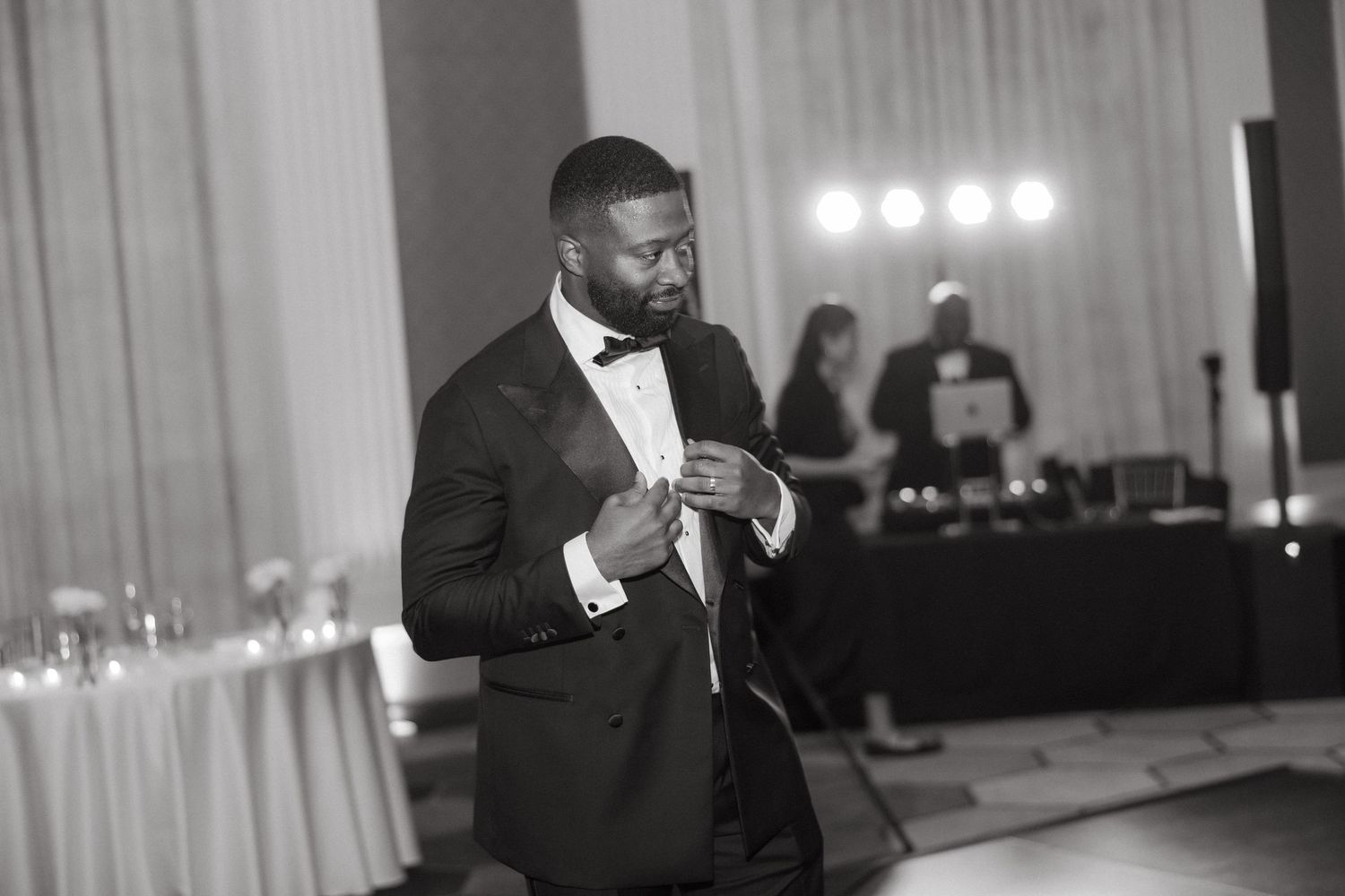 Elegant man in tuxedo and bow tie adjusting cufflinks at a formal black and white wedding reception.