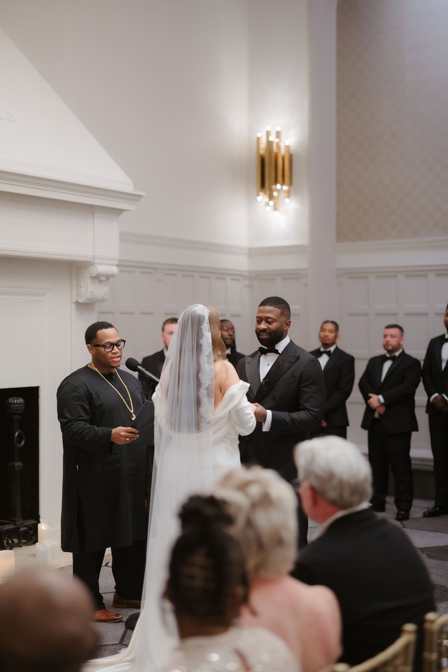 Bride and groom exchange vows at elegant indoor wedding ceremony with officiant and groomsmen in black tuxedos.