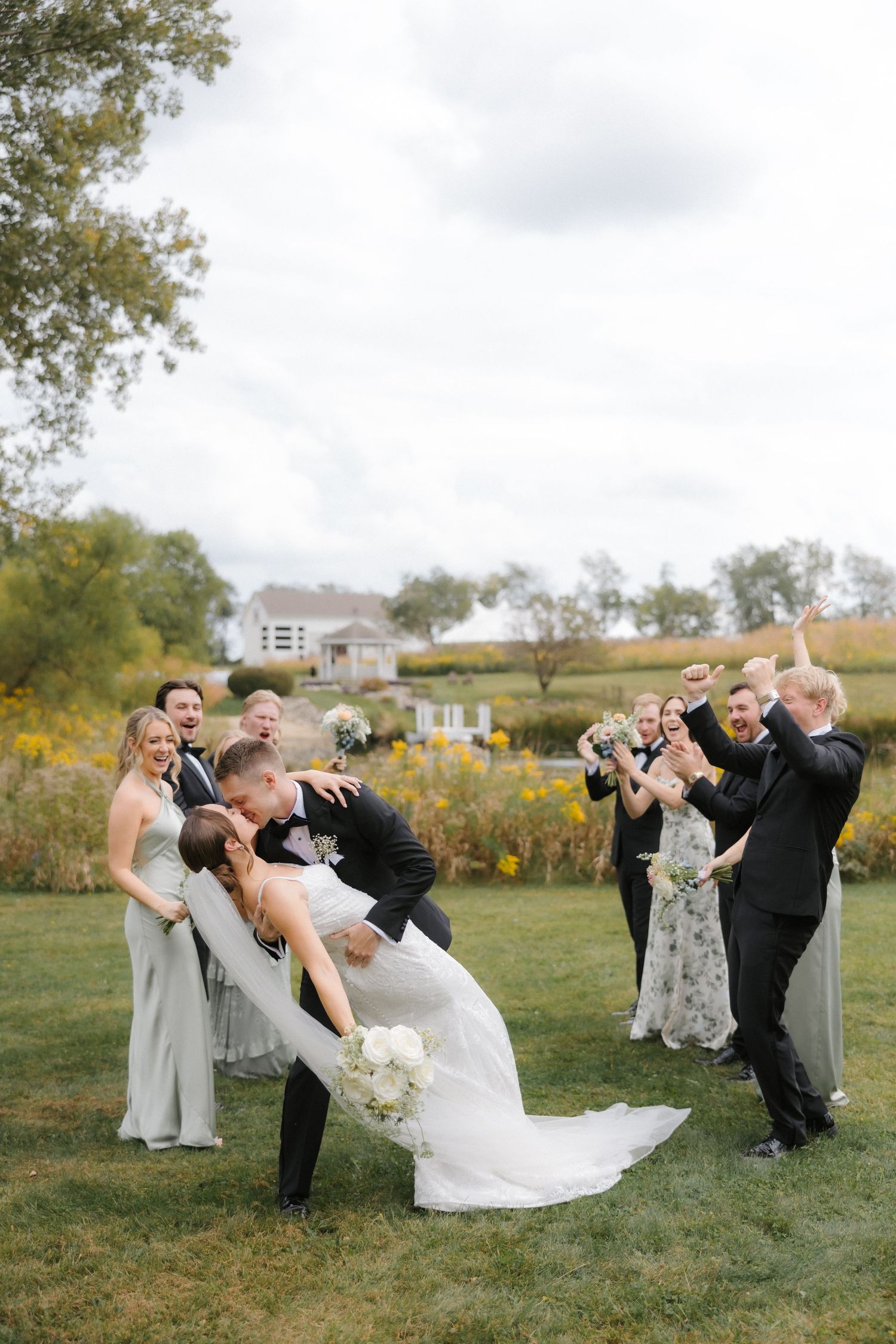 Groom dips bride for a kiss as excited wedding party celebrates with raised bouquets in an autumn outdoor setting.