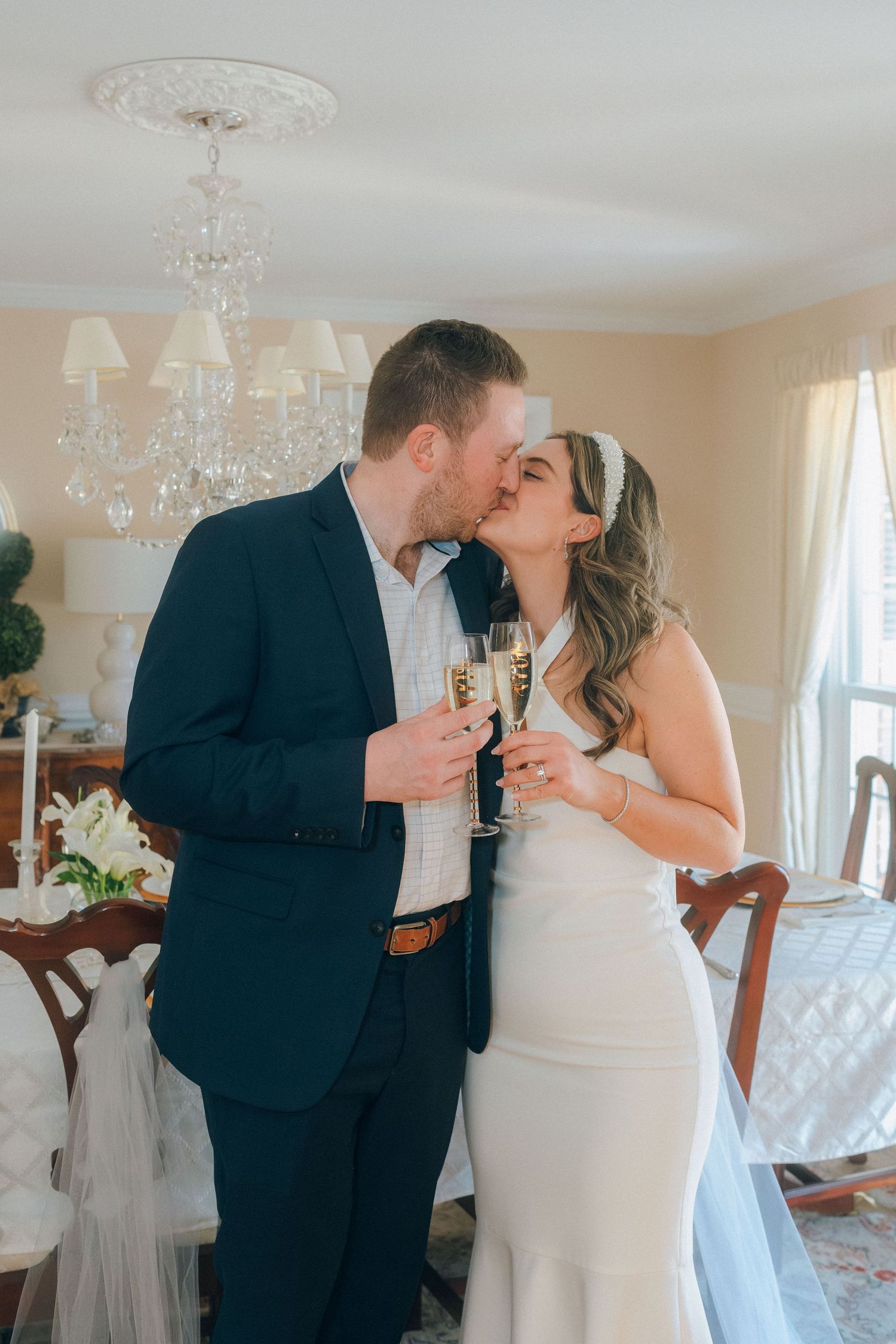 Newlyweds sharing a kiss while holding champagne flutes in an elegant chandelier-lit dining room.