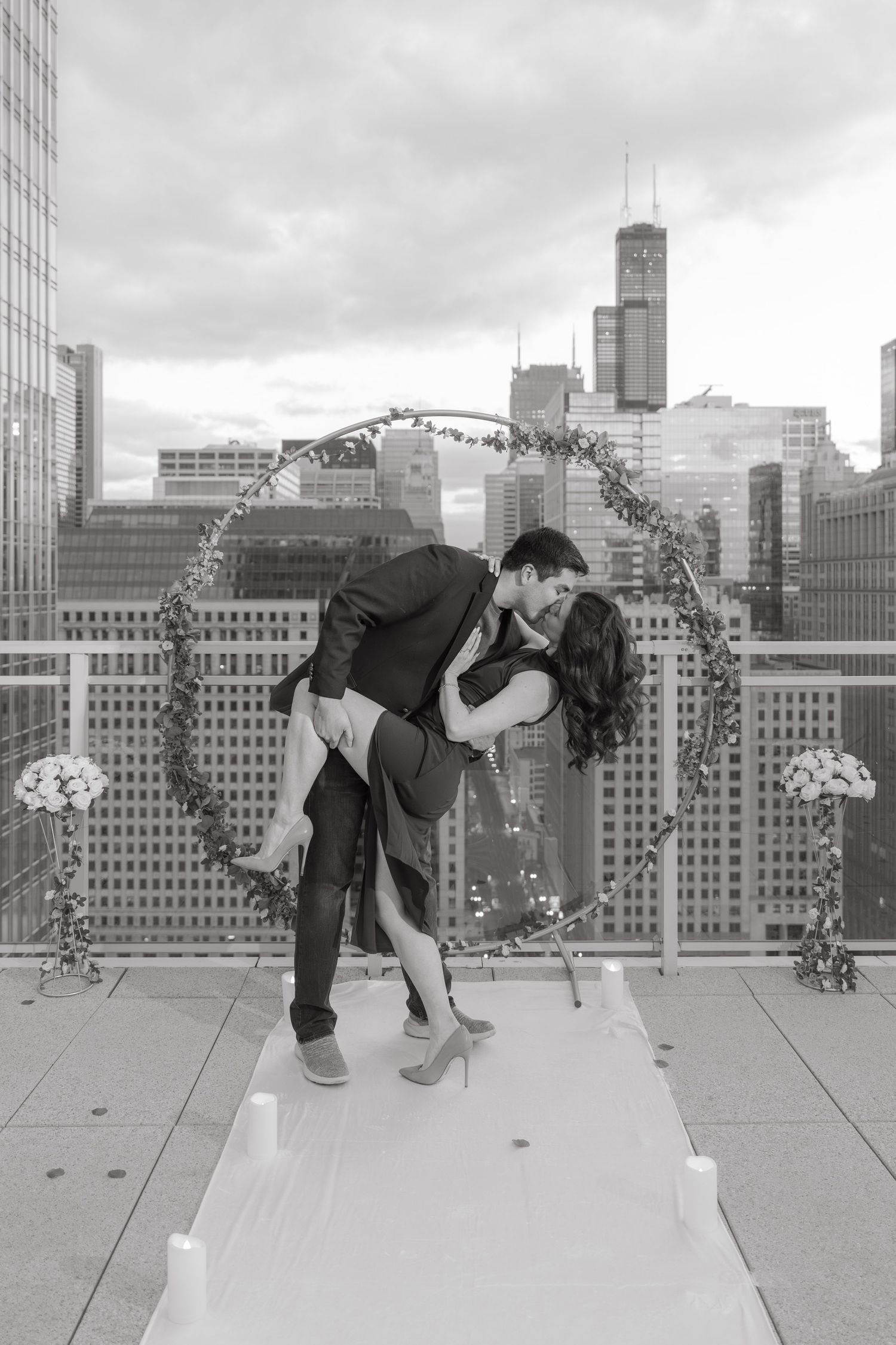 Romantic black and white photo of couple kissing on Chicago rooftop with floral arch and city skyline backdrop.