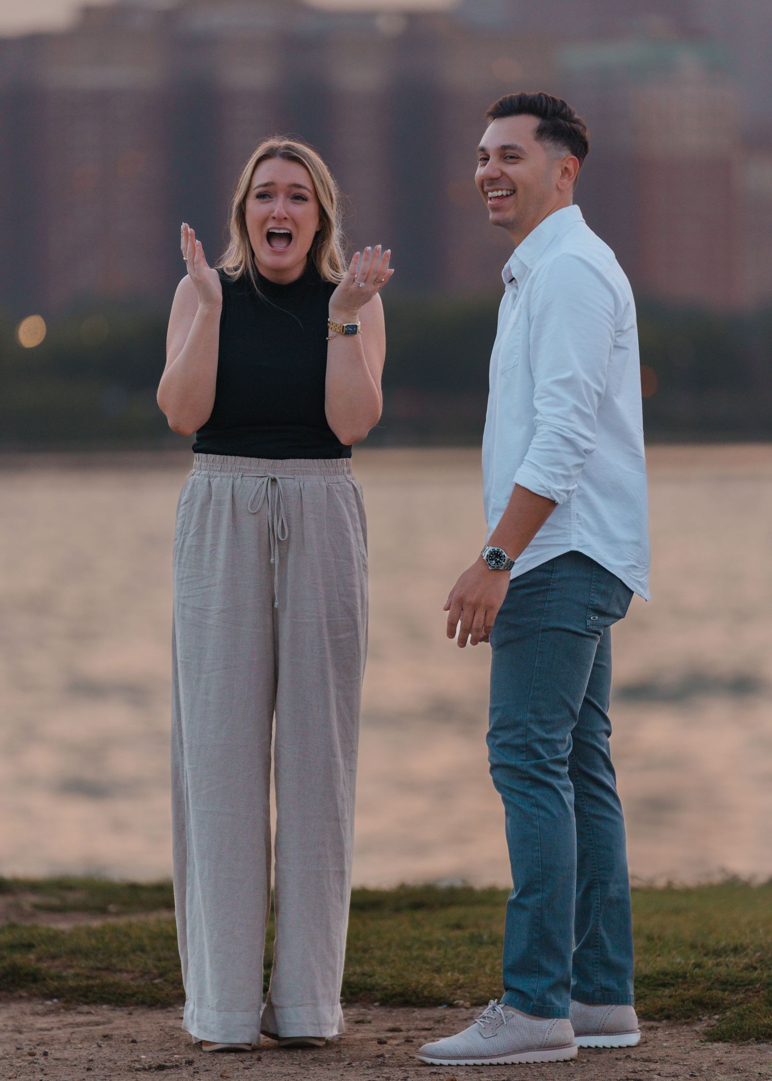 Excited woman clapping hands with laughing man standing by a river at golden hour.