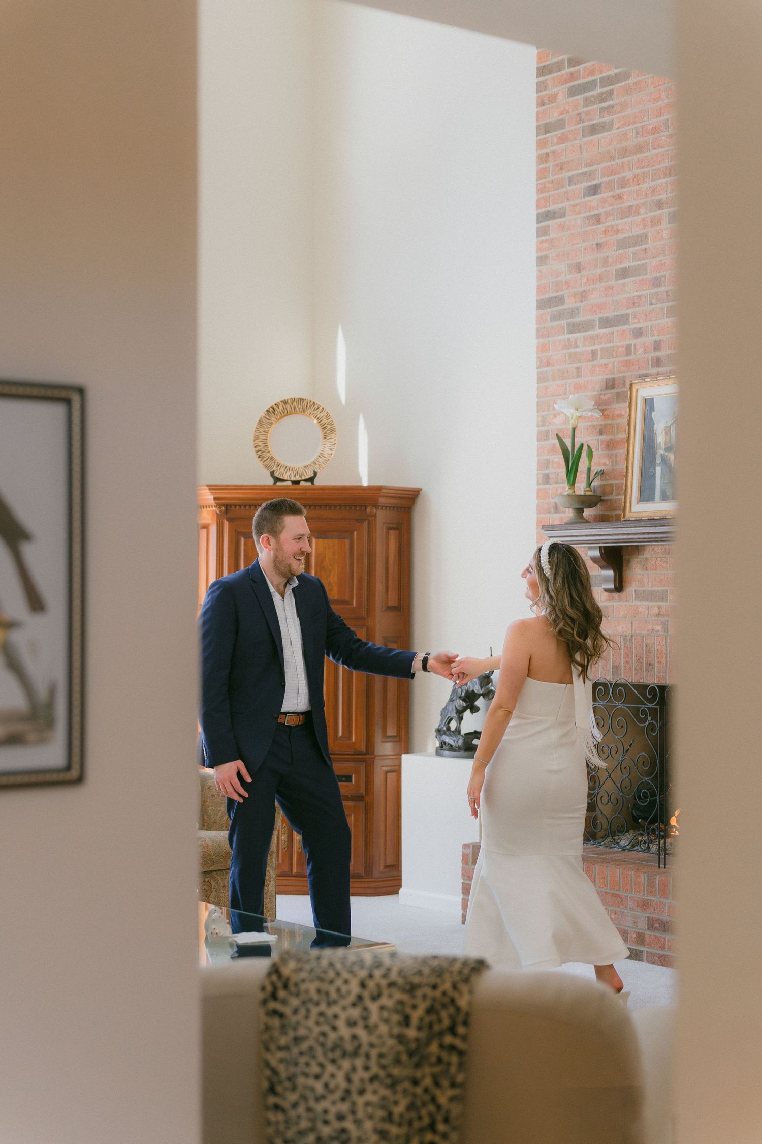 Groom in navy suit holds bride's hand as she twirls in white dress by elegant brick fireplace.