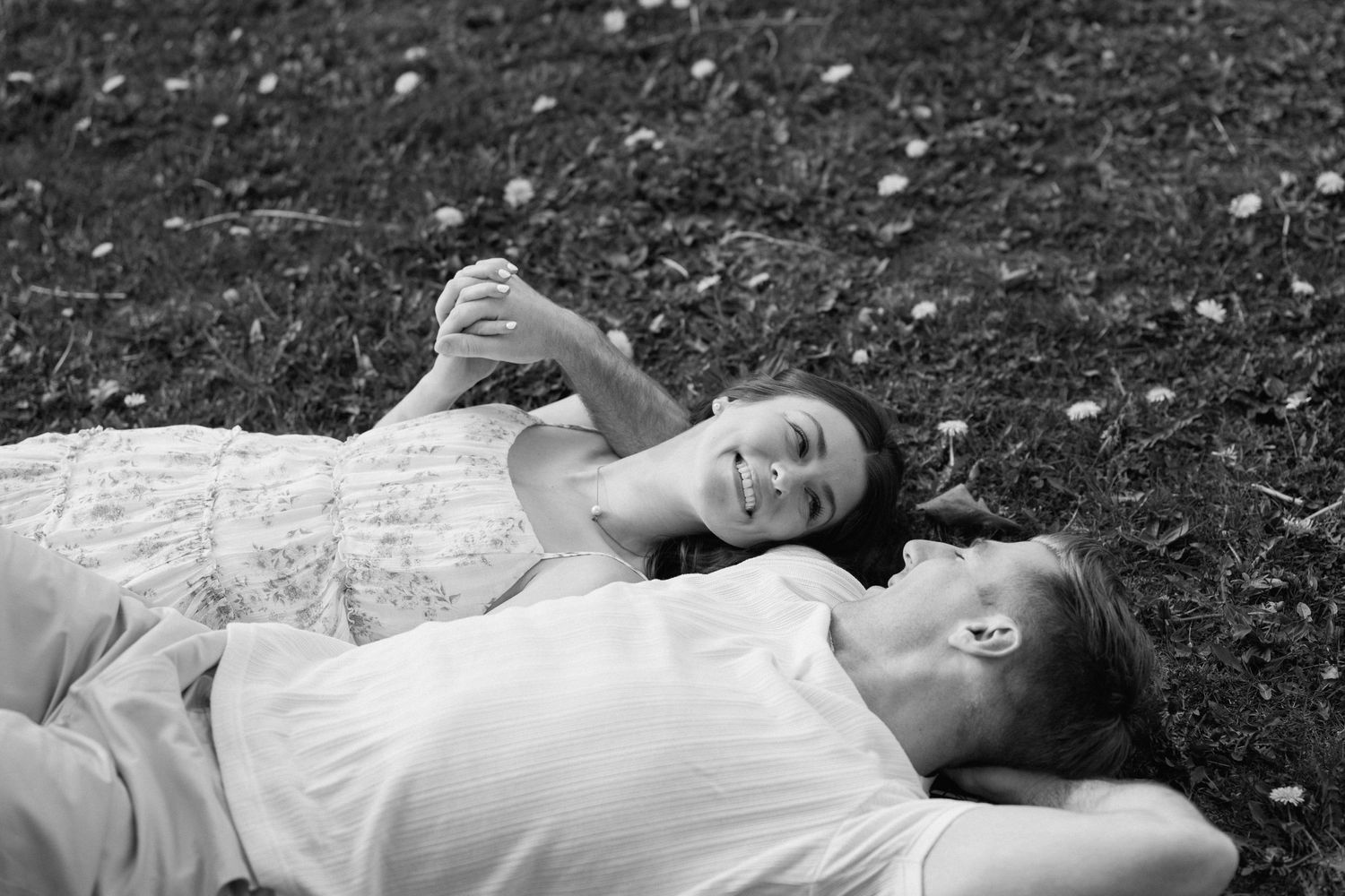Black and white photo of a couple lying in grass holding hands and smiling.