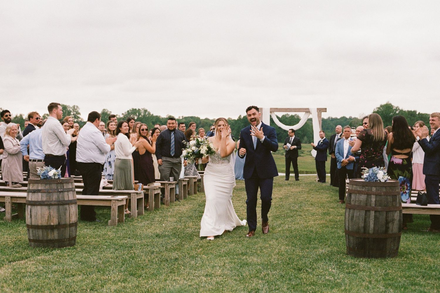 Newlywed couple walks down outdoor wedding aisle lined with wooden benches and barrel decorations on a grassy lawn.