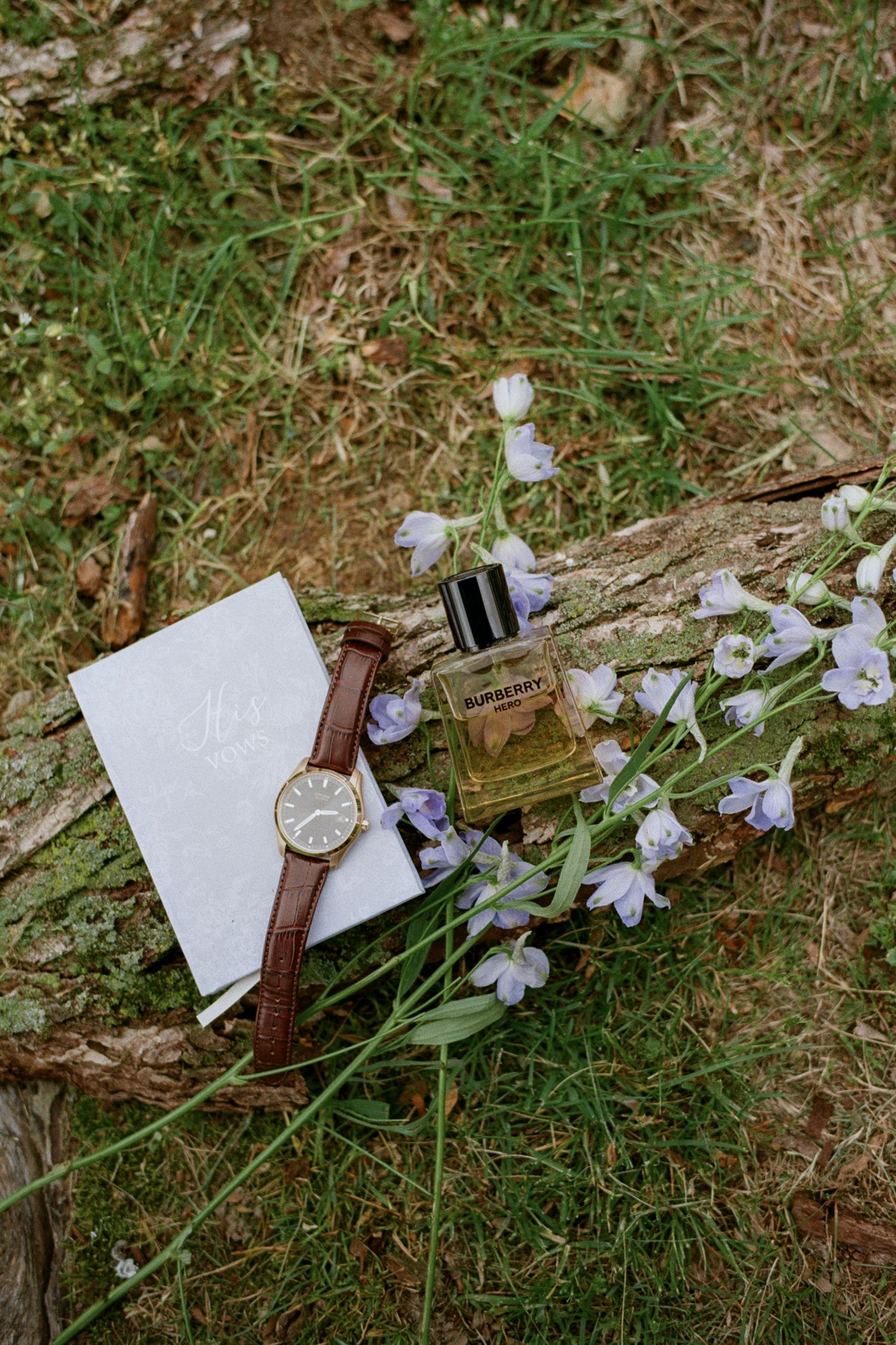Burberry cologne bottle, brown leather watch, and white card on a log with purple wildflowers and green grass.