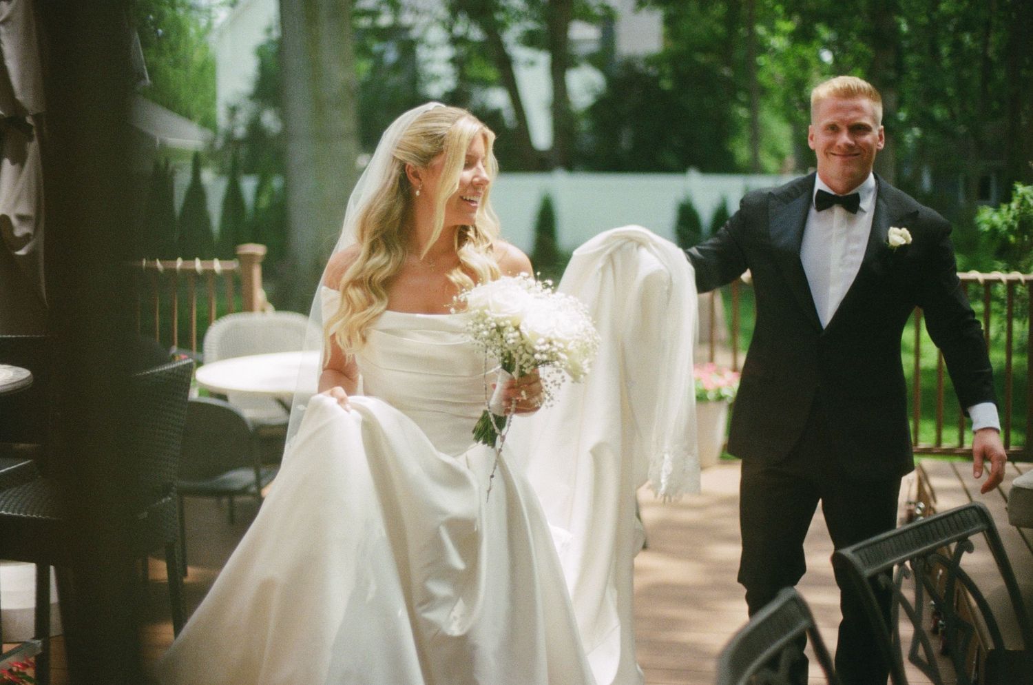 Bride in white gown holding bouquet walks with groom in black tuxedo at outdoor wedding venue.