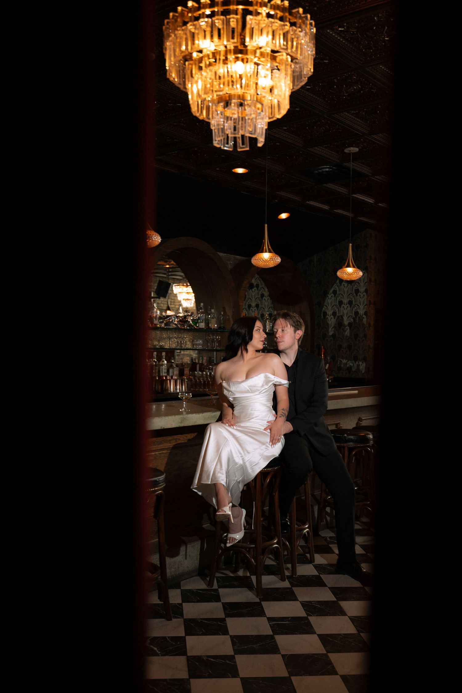 Bride in white satin dress sits at a dark bar with a chandelier overhead as groom in black suit leans in close.