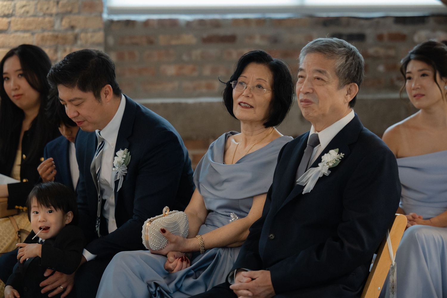 Elderly Asian couple in formal attire sitting at a wedding ceremony, with family members including a young child nearby.