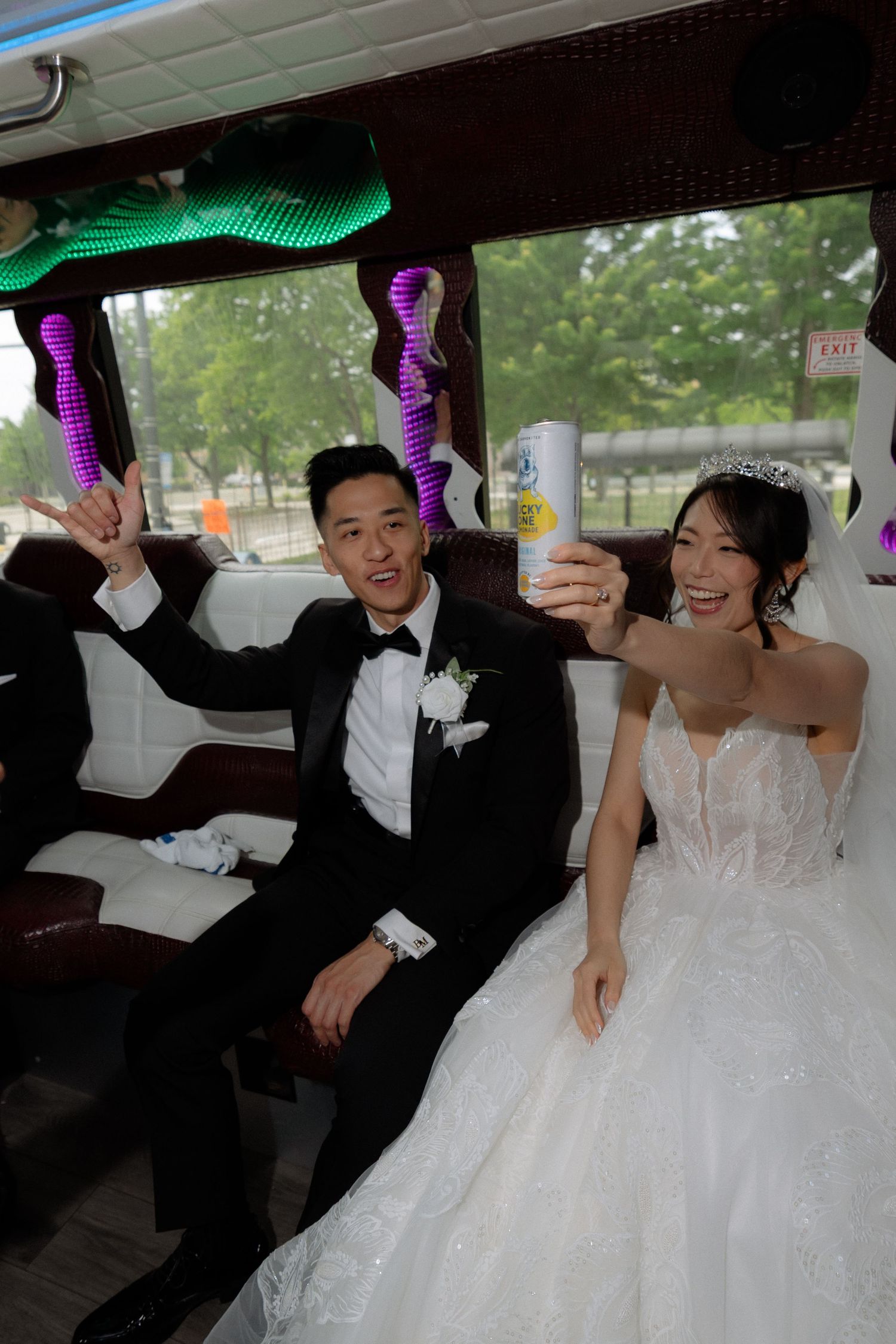 Happy Asian bride and groom celebrating in decorated party bus holding drinks on wedding day.
