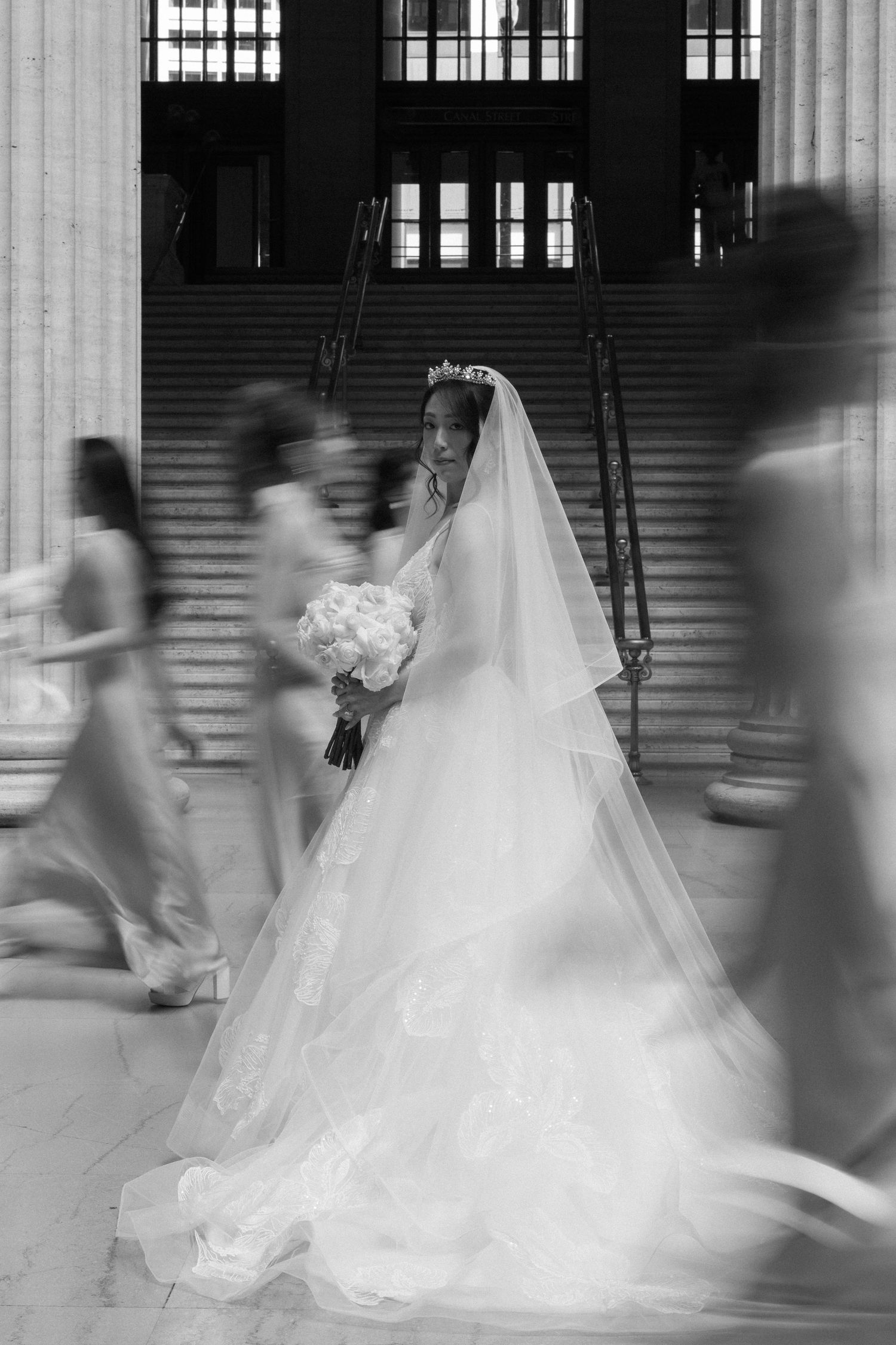 Bride in elegant white gown and veil holds bouquet on grand staircase in black and white photo.