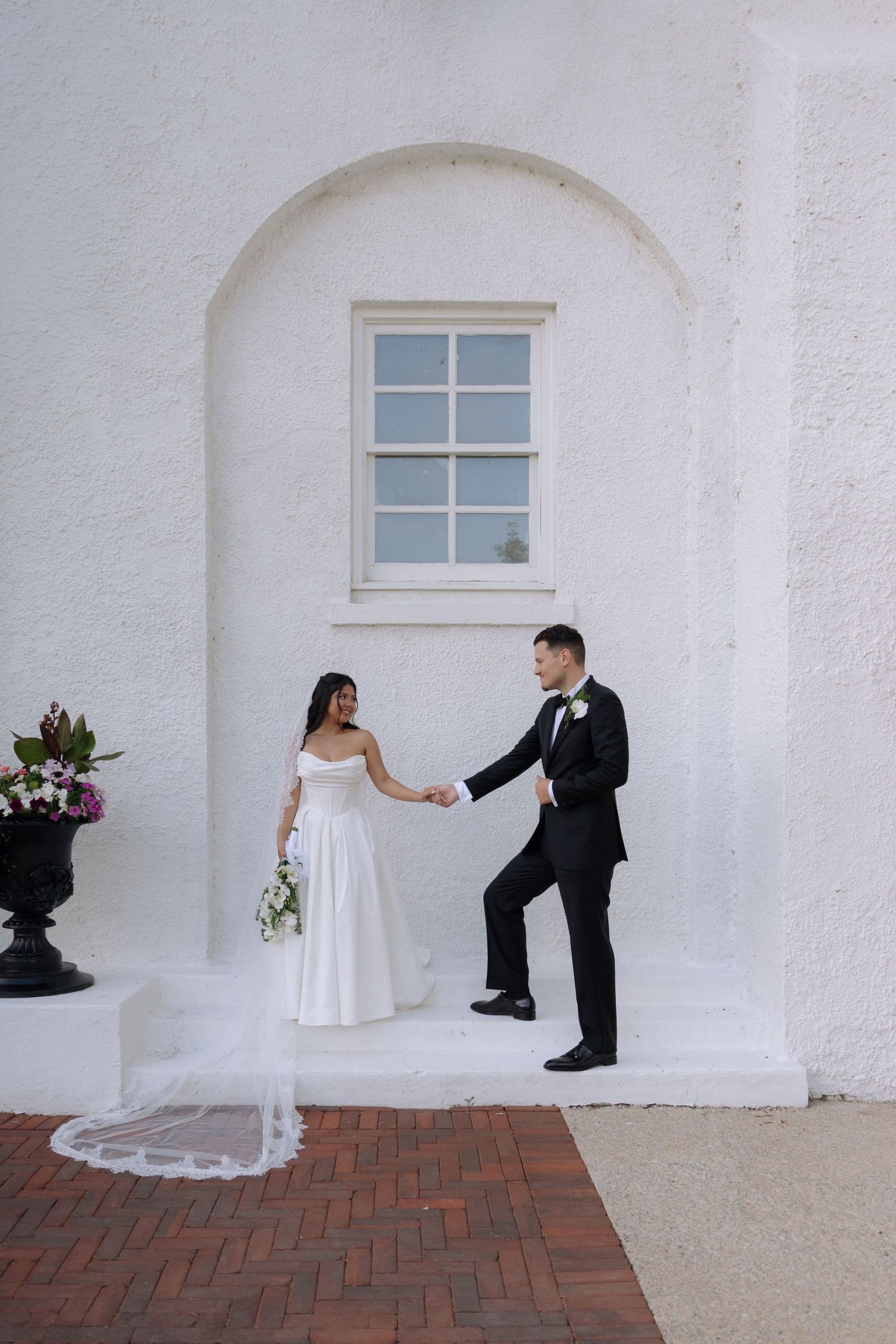 Bride and groom hold hands on brick steps against a white arched wall, bride in elegant gown with veil.