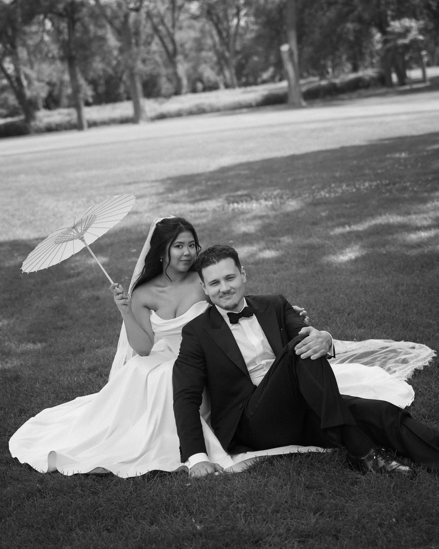 Black and white photo of bride and groom sitting on grass, bride holding white parasol, groom in tuxedo.
