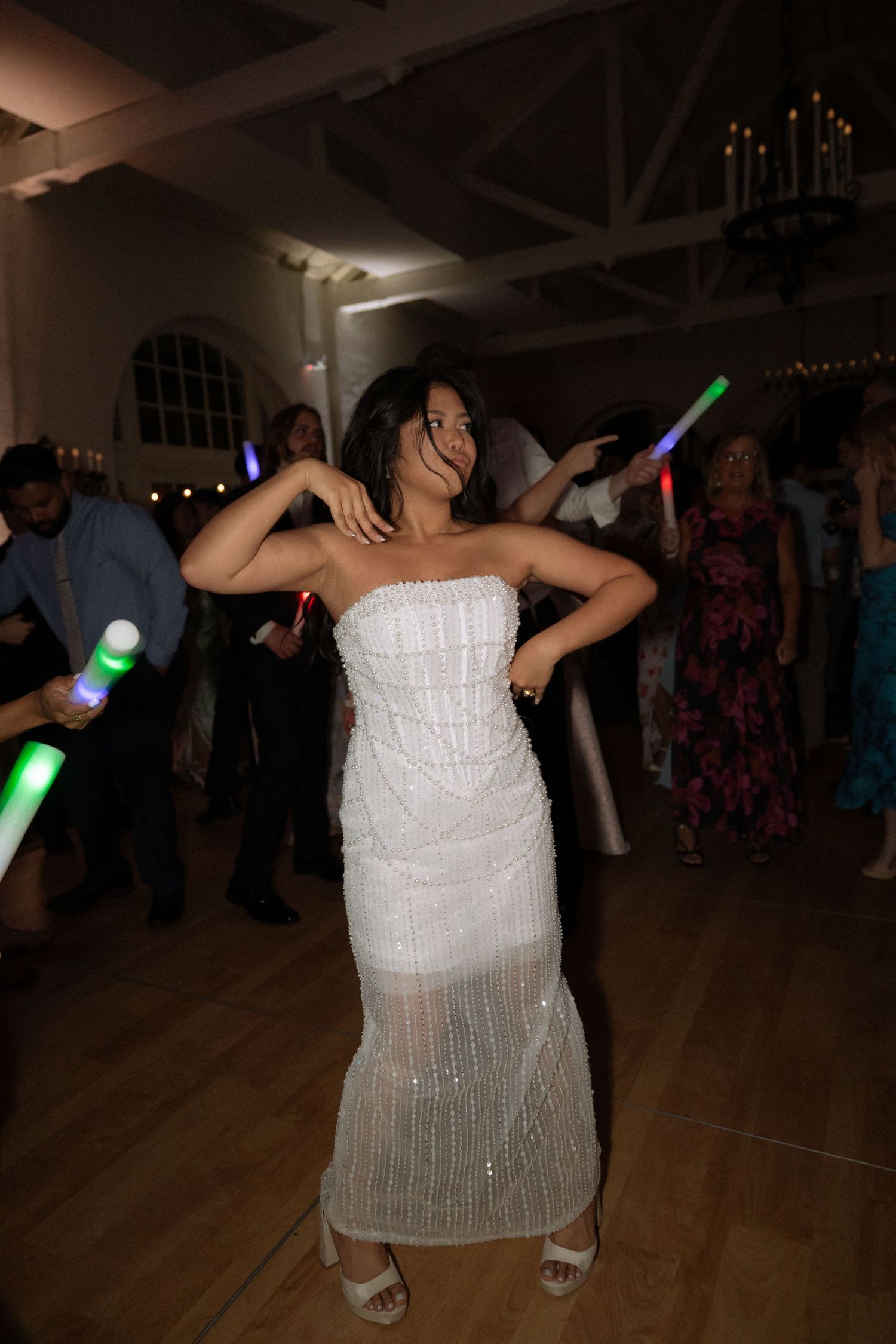 Bride in white strapless gown dancing joyfully at wedding reception with glowing light sticks in background.