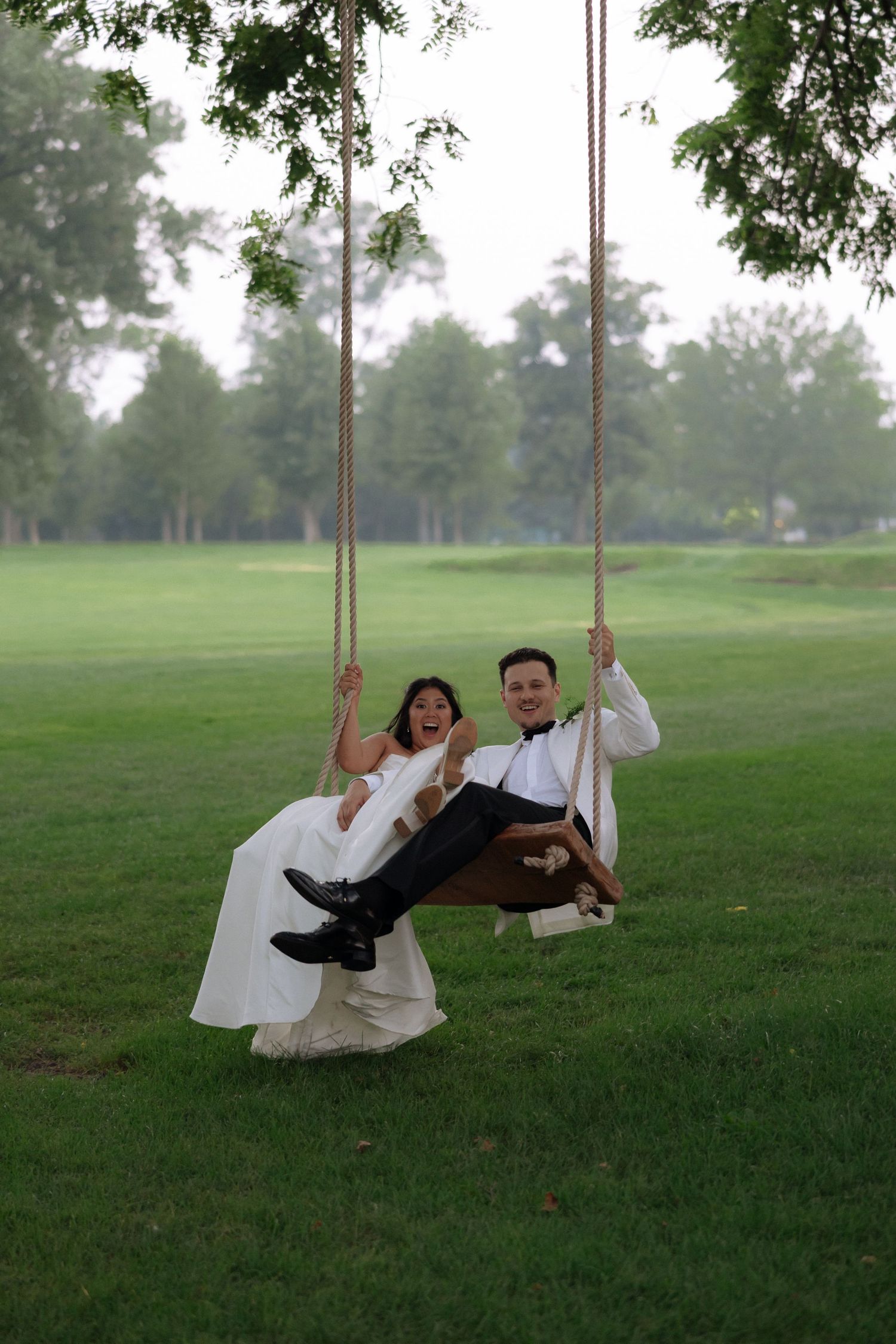 Joyful bride and groom sharing a wooden rope swing in a lush green park on their wedding day.
