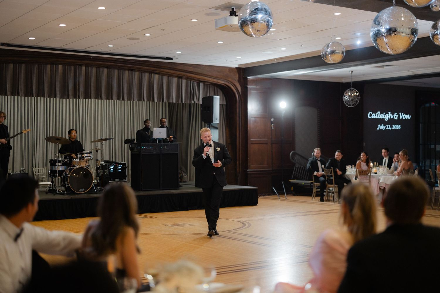 Wedding MC speaking at microphone during elegant reception with disco balls and personalized backdrop display.