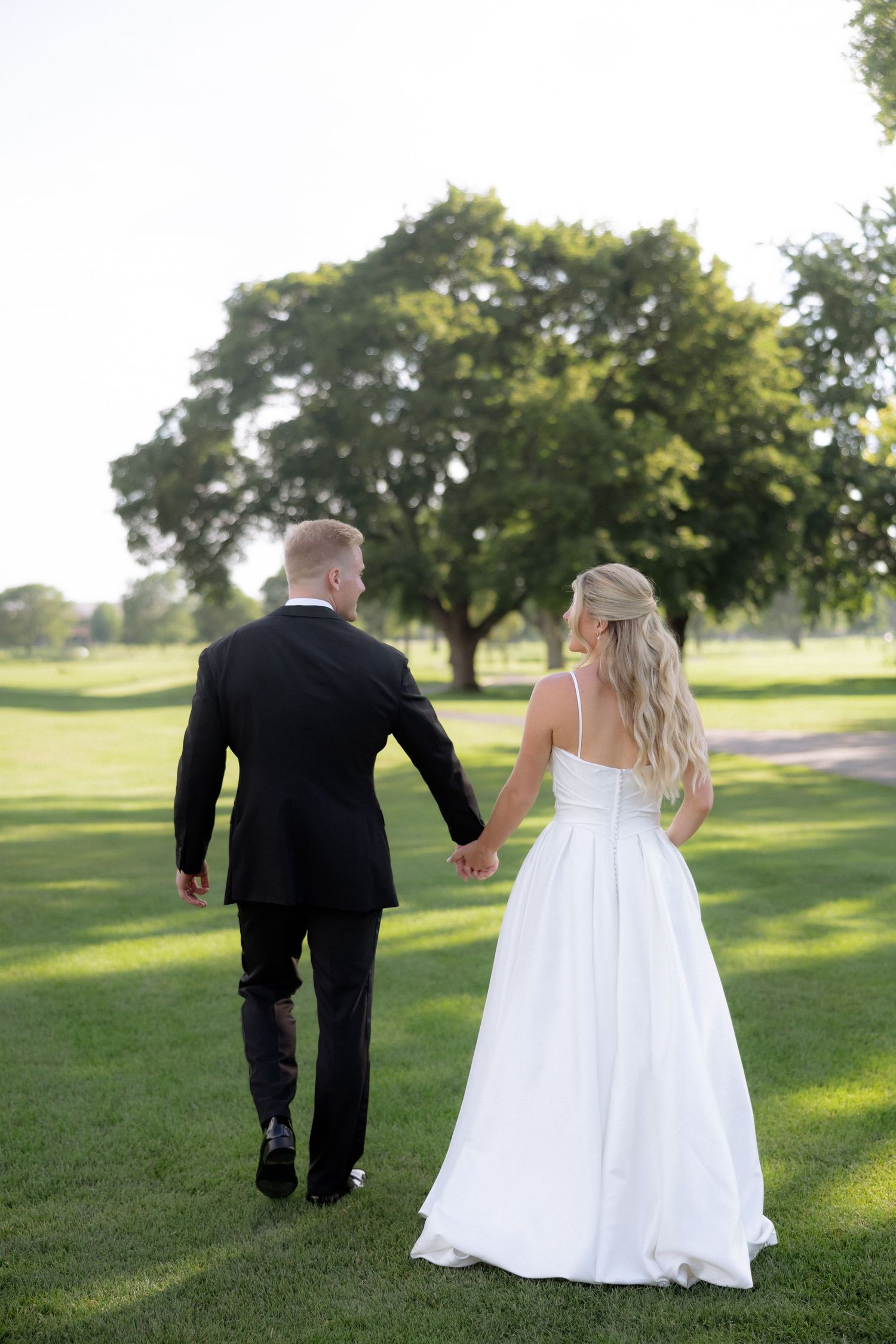 Bride and groom walk hand in hand across lush green lawn surrounded by trees in golden light.