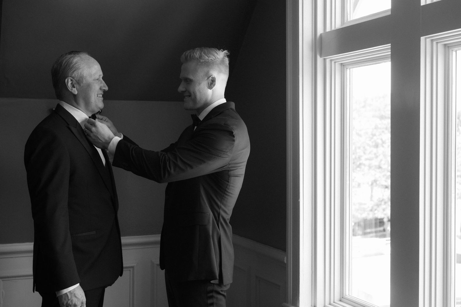 Groom adjusts older mans tie near window in elegant black and white wedding preparation photo.