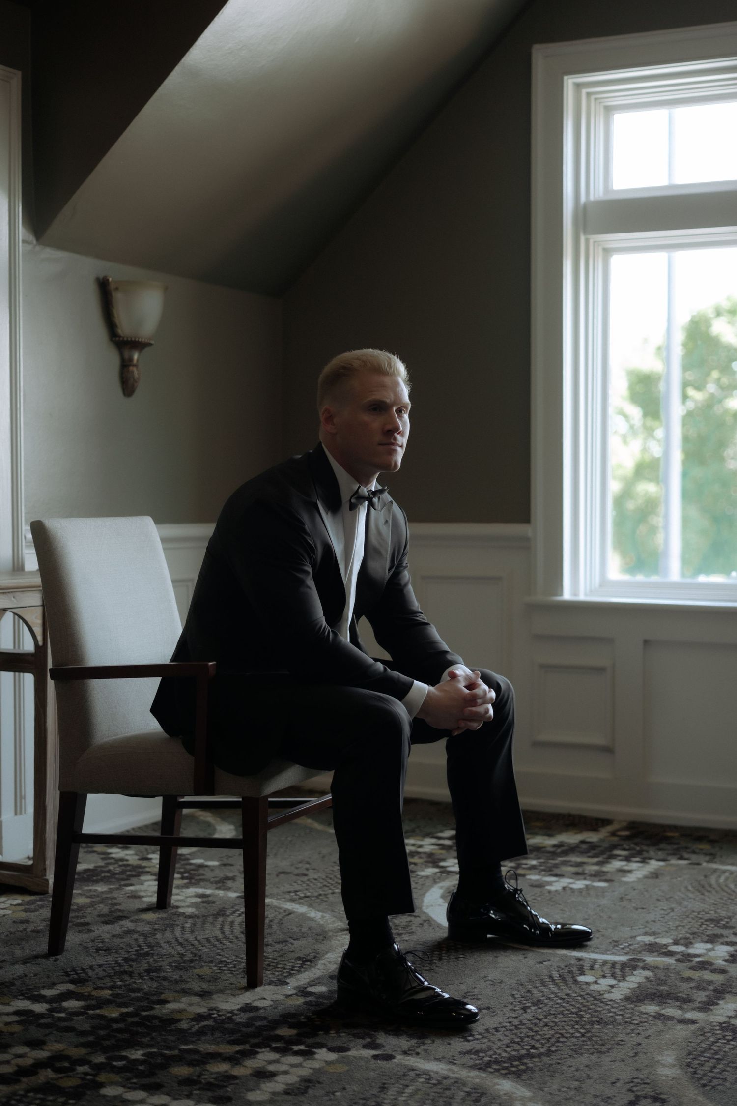 Groom in black tuxedo with bow tie sitting thoughtfully in a chair by a window before the wedding.