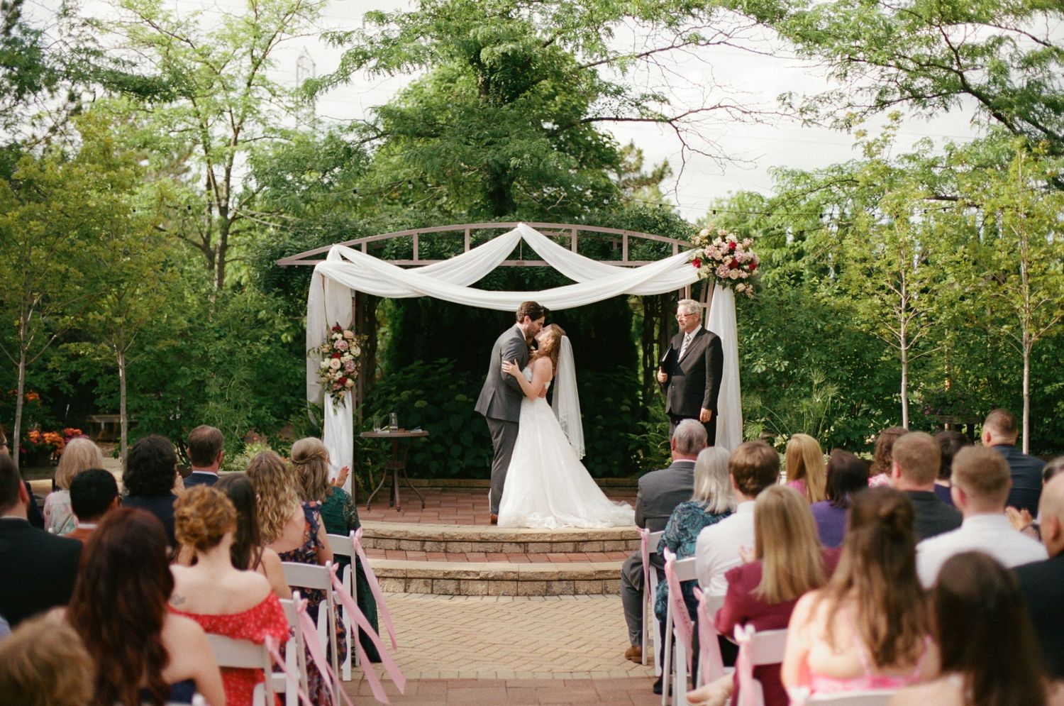 Bride and groom kiss at outdoor wedding ceremony under floral arch, surrounded by seated guests and lush green trees.