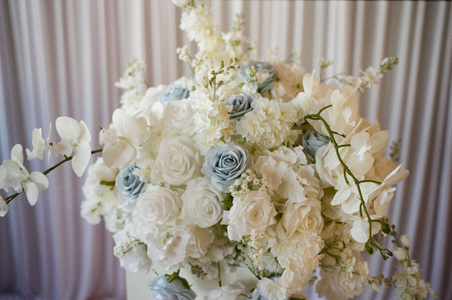 Elegant wedding floral arrangement featuring white roses, blue roses, orchids, and delicate white blooms against a soft draped backdrop.