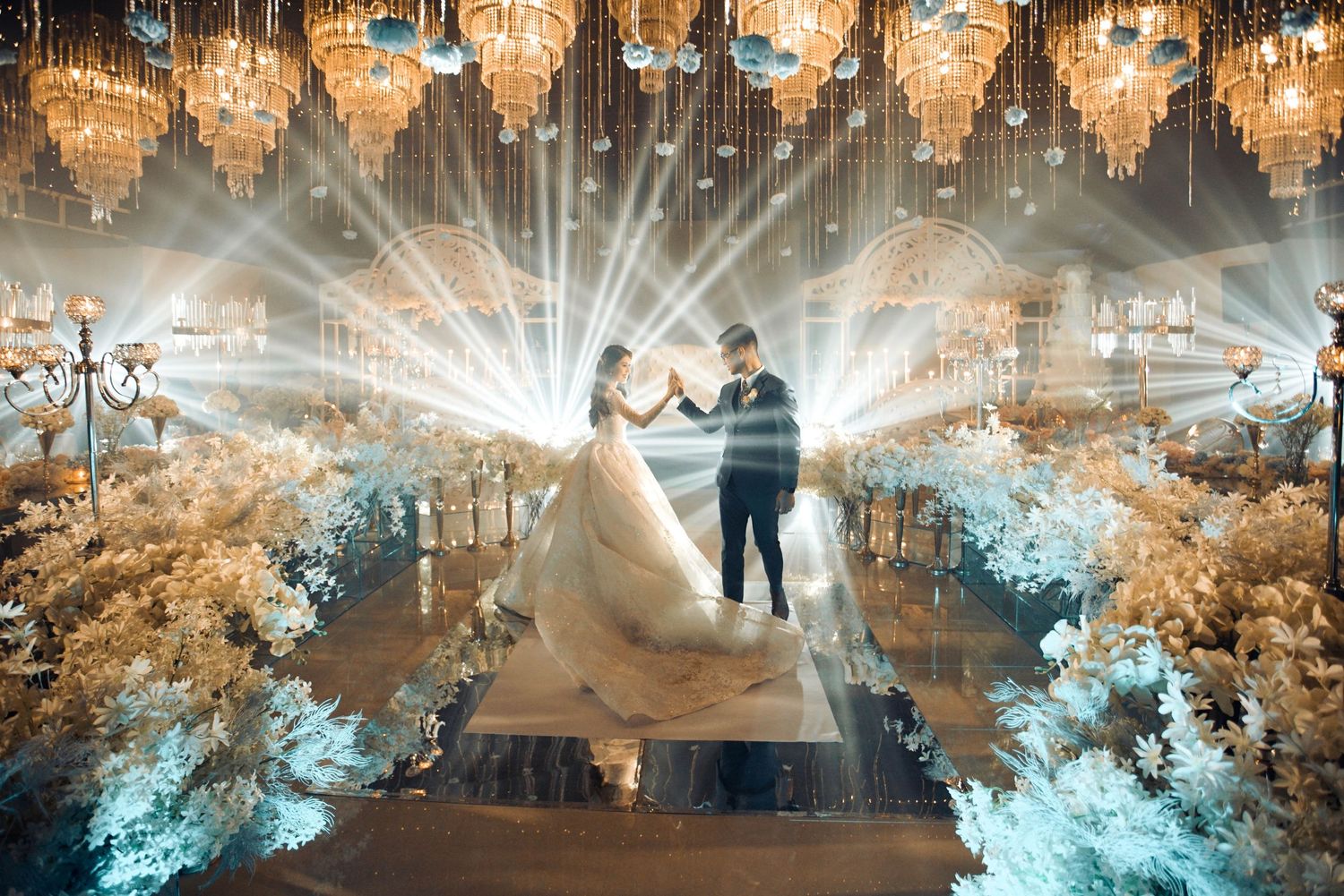 Couple dancing at elegant wedding reception with crystal chandeliers, dramatic lighting, and white floral arrangements lining the aisle.