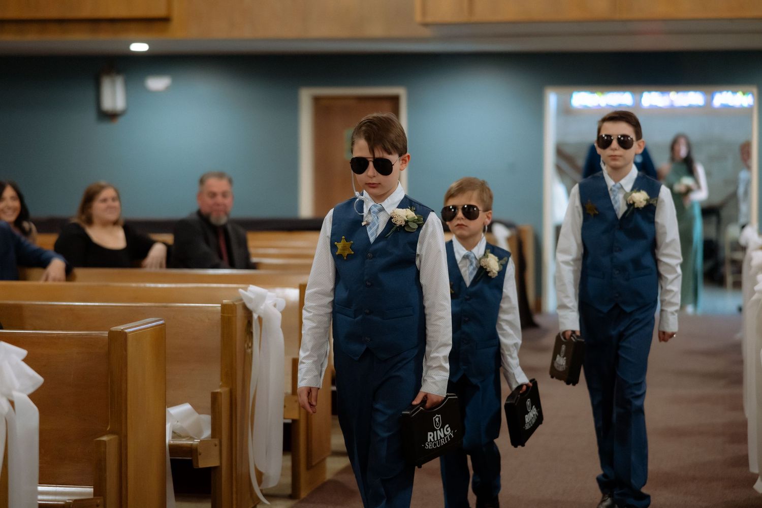 Three young ring bearers in navy suits and sunglasses walk down a church aisle during a wedding ceremony.