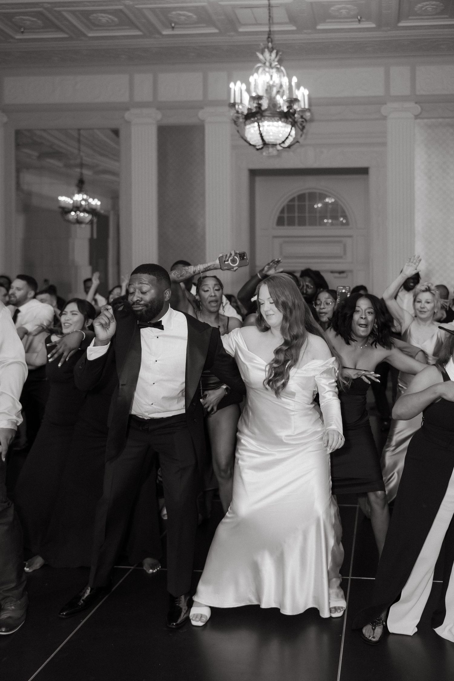 Black and white photo of a bride and groom dancing joyfully at their elegant wedding reception with guests celebrating behind them.