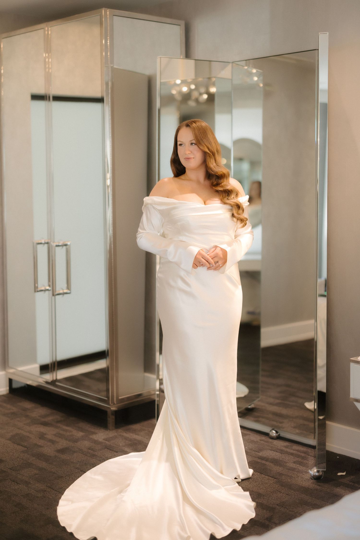 Bride wearing an elegant off-shoulder white satin wedding gown with long sleeves and a train in a bridal boutique.