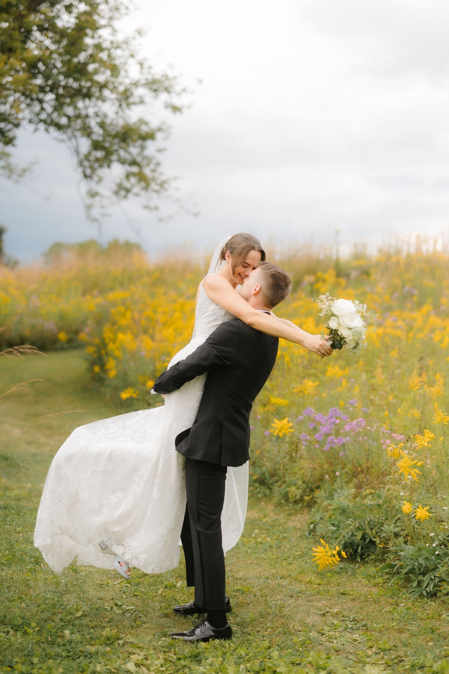 Groom lifting bride in white gown and veil in front of a colorful wildflower field.