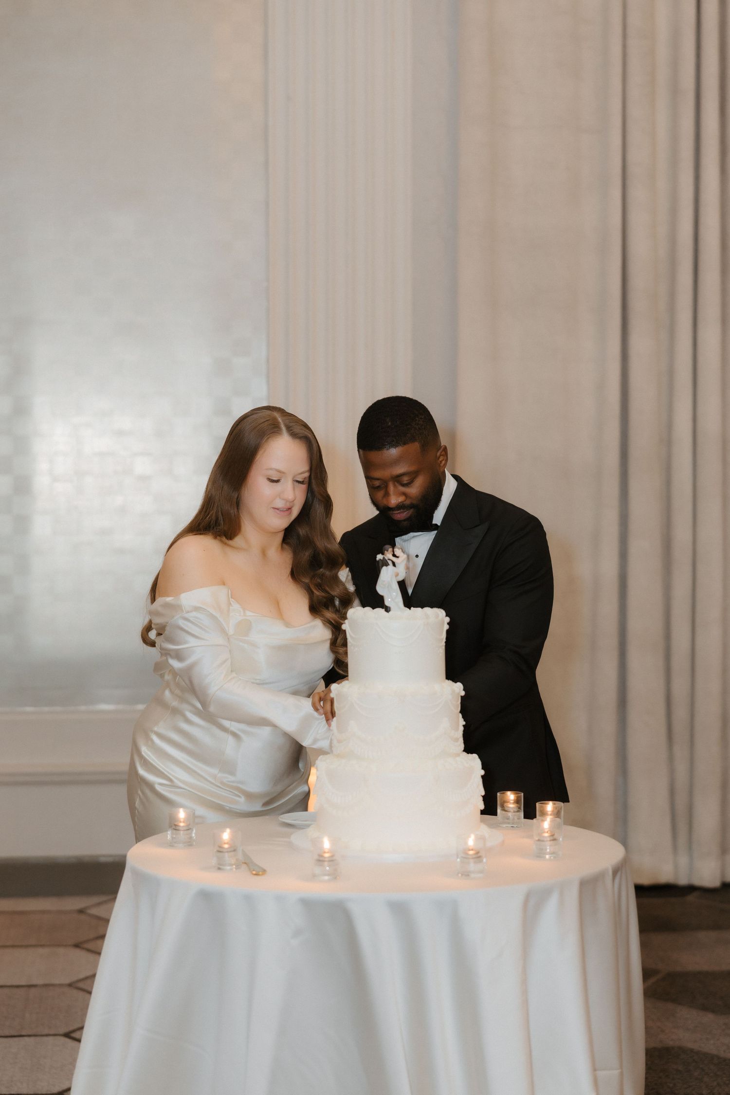 Bride and groom cutting a white tiered wedding cake at a candlelit table in an elegant ballroom.