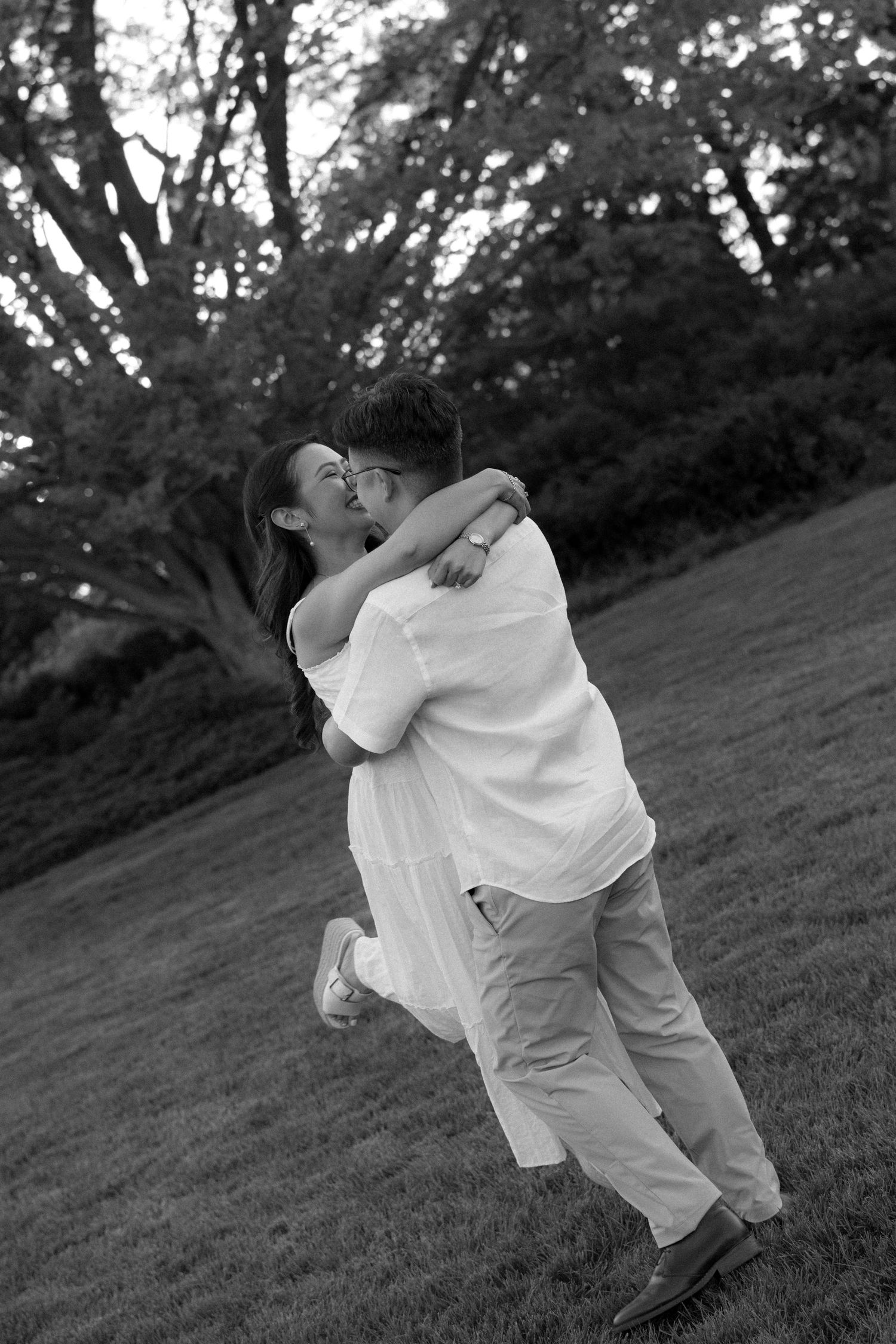 Black and white photo of a couple embracing and kissing in a park, surrounded by lush trees and green grass.