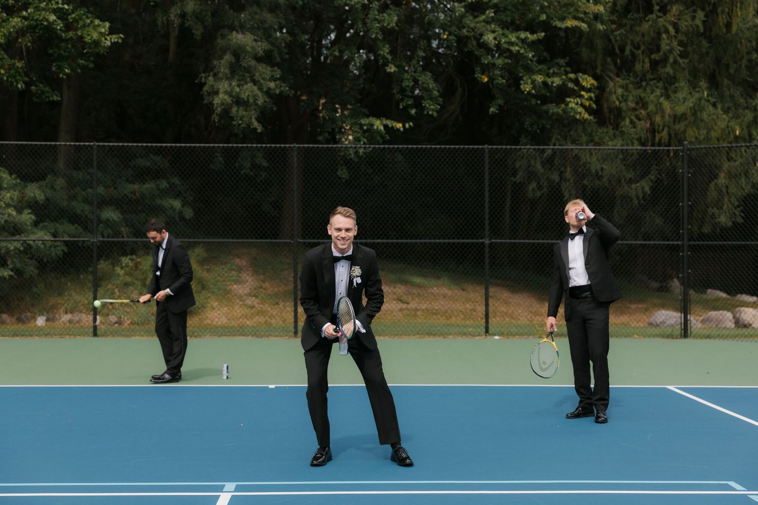 Groomsmen in black tuxedos celebrate on a blue tennis court, with one dancing joyfully in the center.