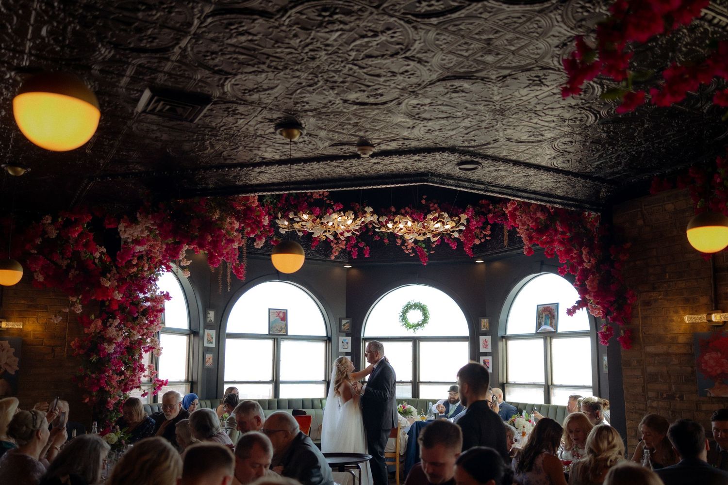 Elegant wedding reception in a vintage venue with red floral arch, arched windows, and guests seated at candlelit tables.